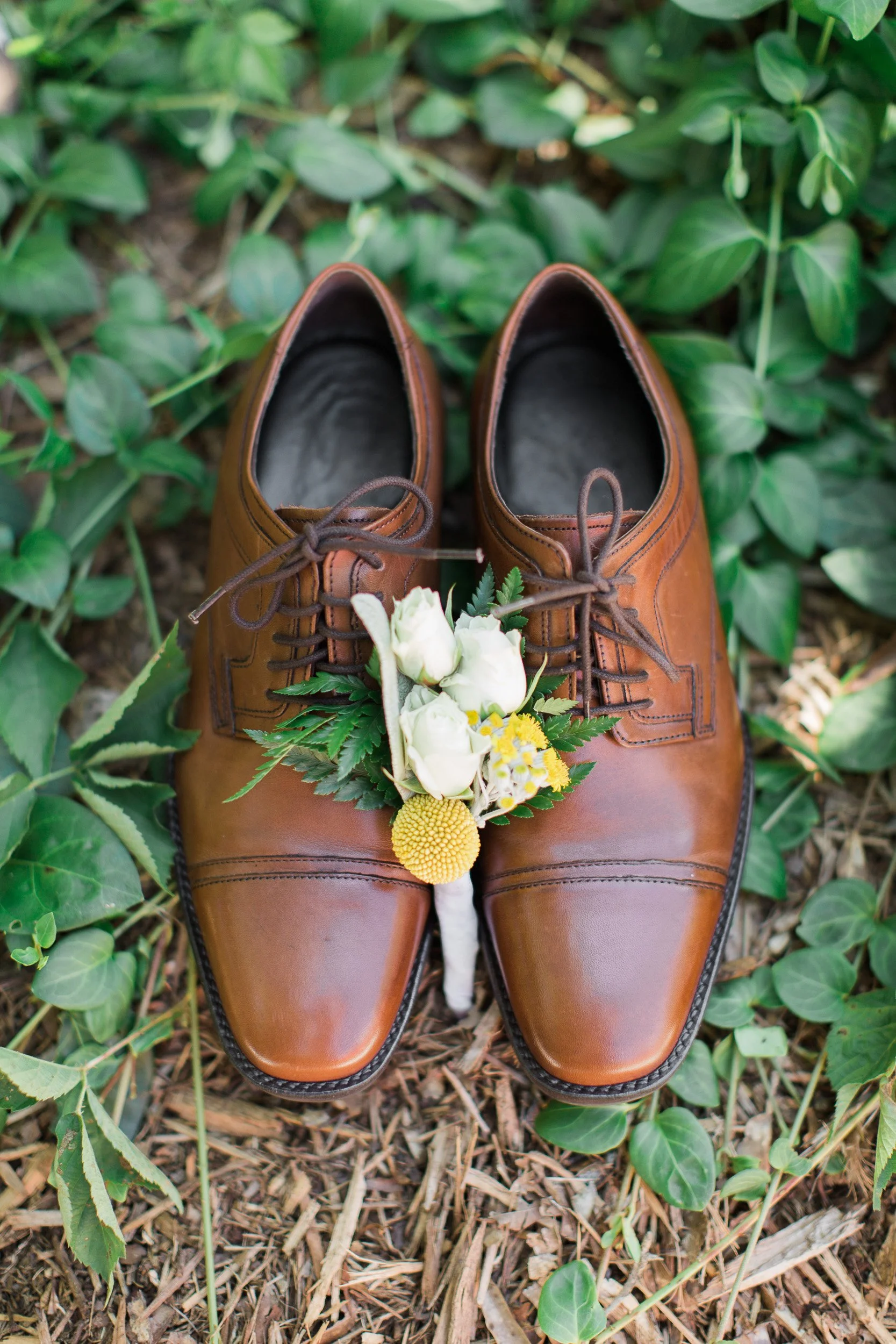 A pair of brown leather dress shoes placed on the ground surrounded by green plants and foliage, with a small bouquet of white and yellow flowers resting on the shoes during backyard wedding in Greenville SC