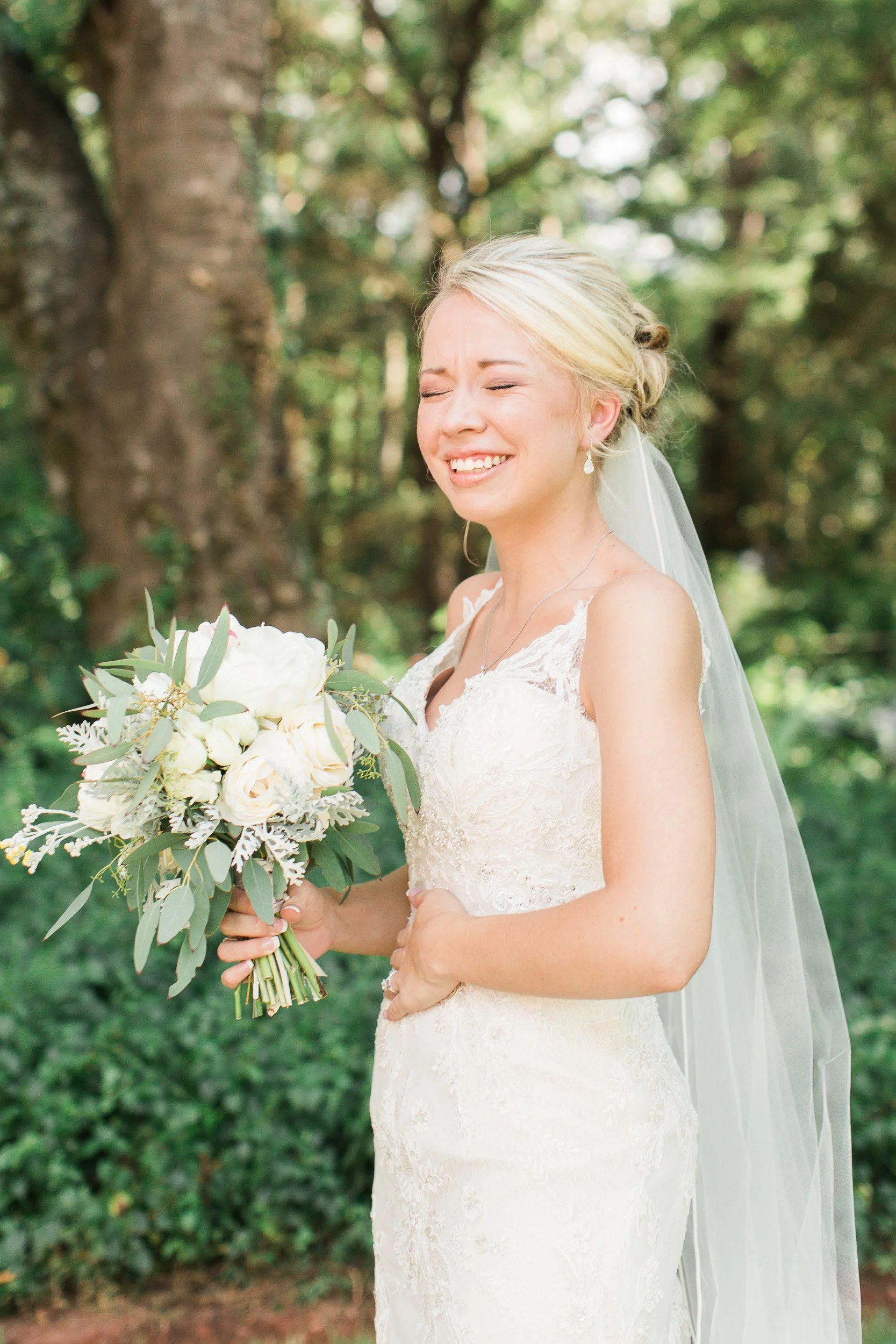 A smiling bride in a lace wedding dress holding a bouquet of white flowers and greenery, with a large tree in the background during backyard wedding in Greenville, SC