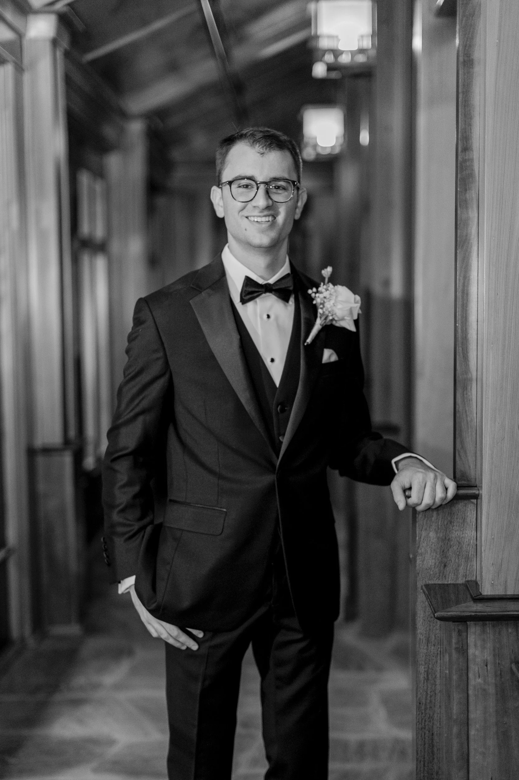 A young man in a tuxedo with a boutonnière, wearing glasses, smiling and posing for intimate wedding in Greenville, SC