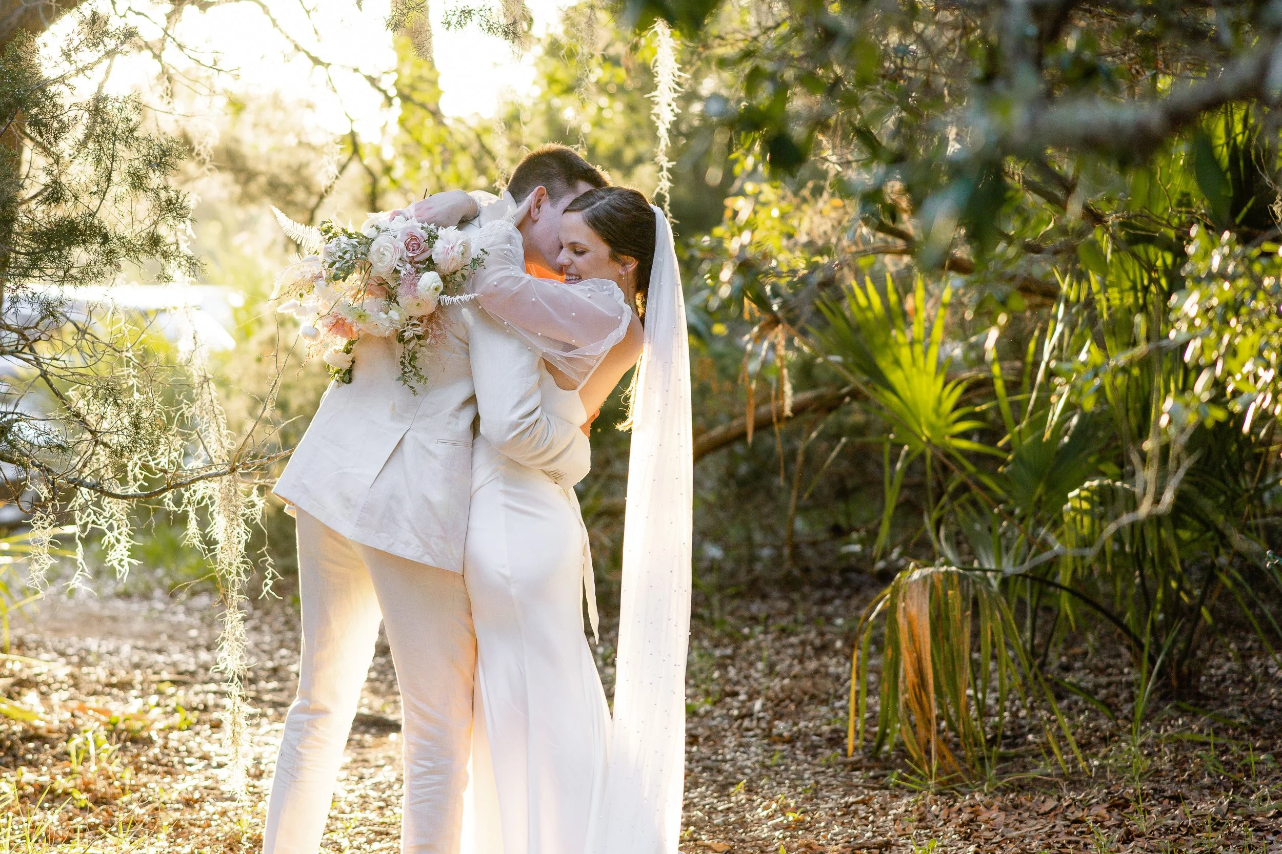 A bride and groom sharing a kiss outdoors, surrounded by greenery and sunlight, with the bride holding a bouquet of flowers during an elopement in Clemson, SC