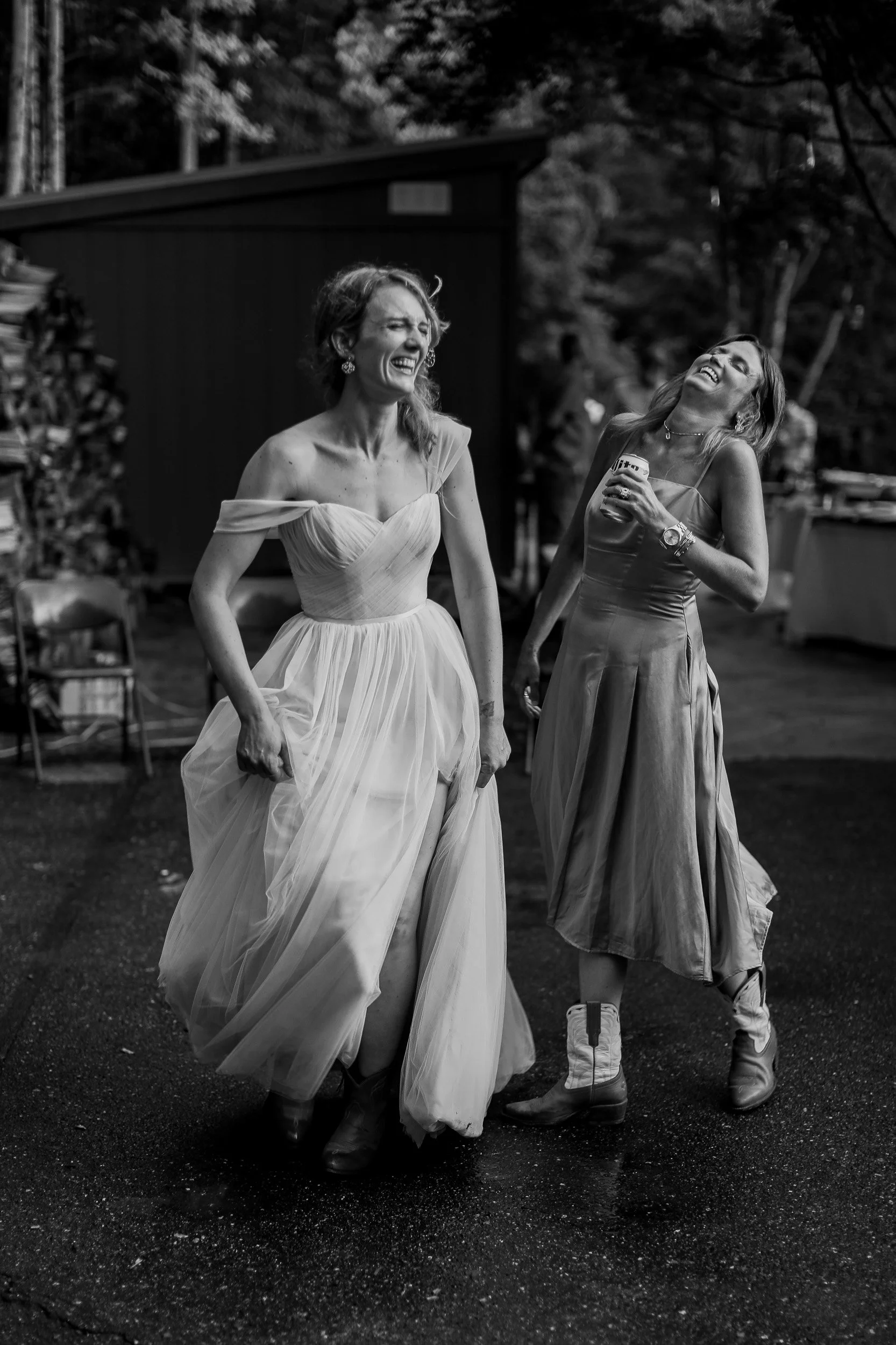 Two women laughing and dancing outdoors at night. One woman wears a light-colored gown, and the other wears a dress with casual boots. They appear to be enjoying themselves.