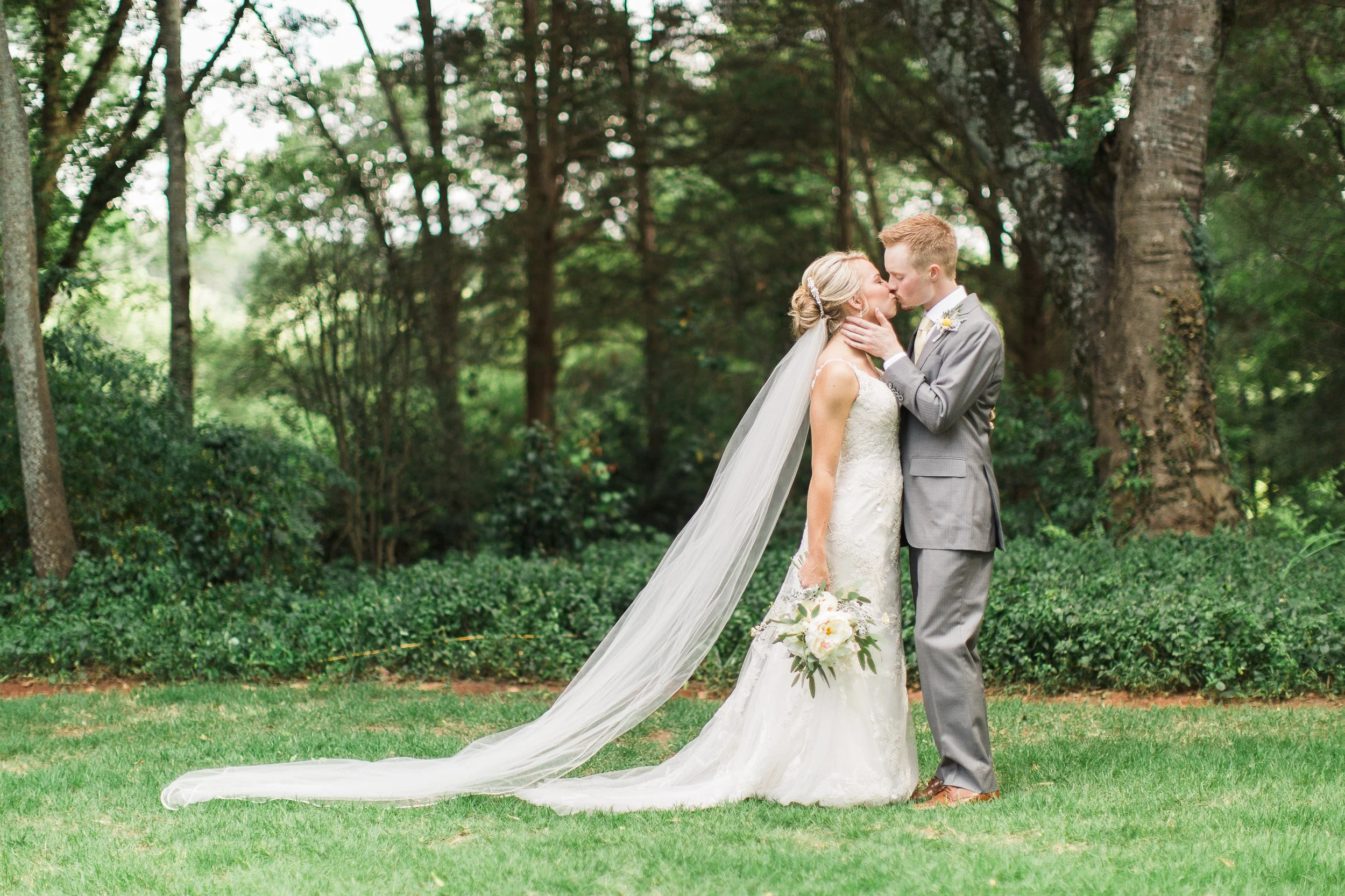 A bride and groom sharing a kiss outdoors during their wedding ceremony, surrounded by green trees and grass at Sleepy Hollow Barn in Pendleton SC