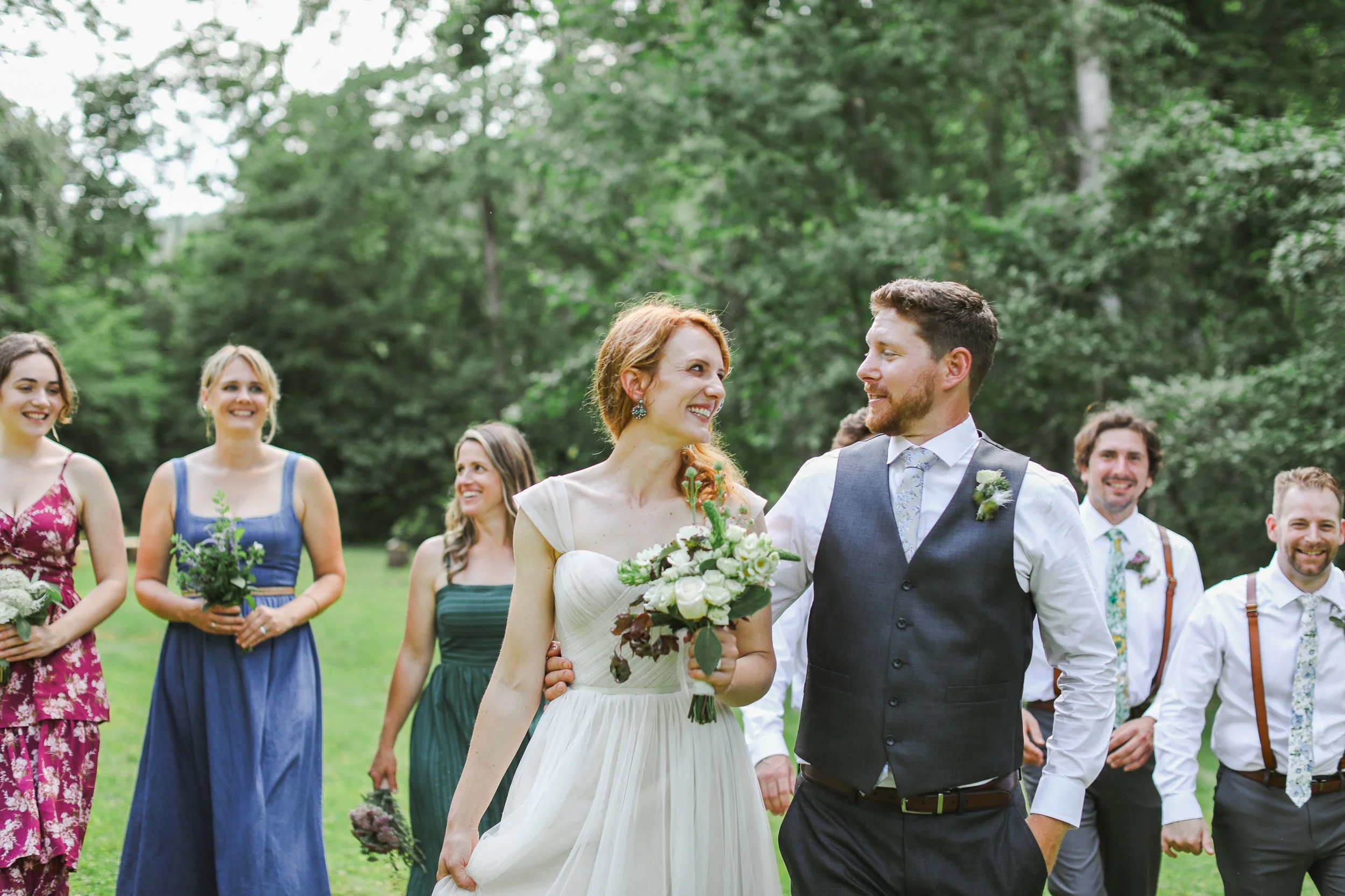 A bride and groom walking hand in hand at their outdoor wedding ceremony, surrounded by bridesmaids and groomsmen, in a lush park in Greenville, SC