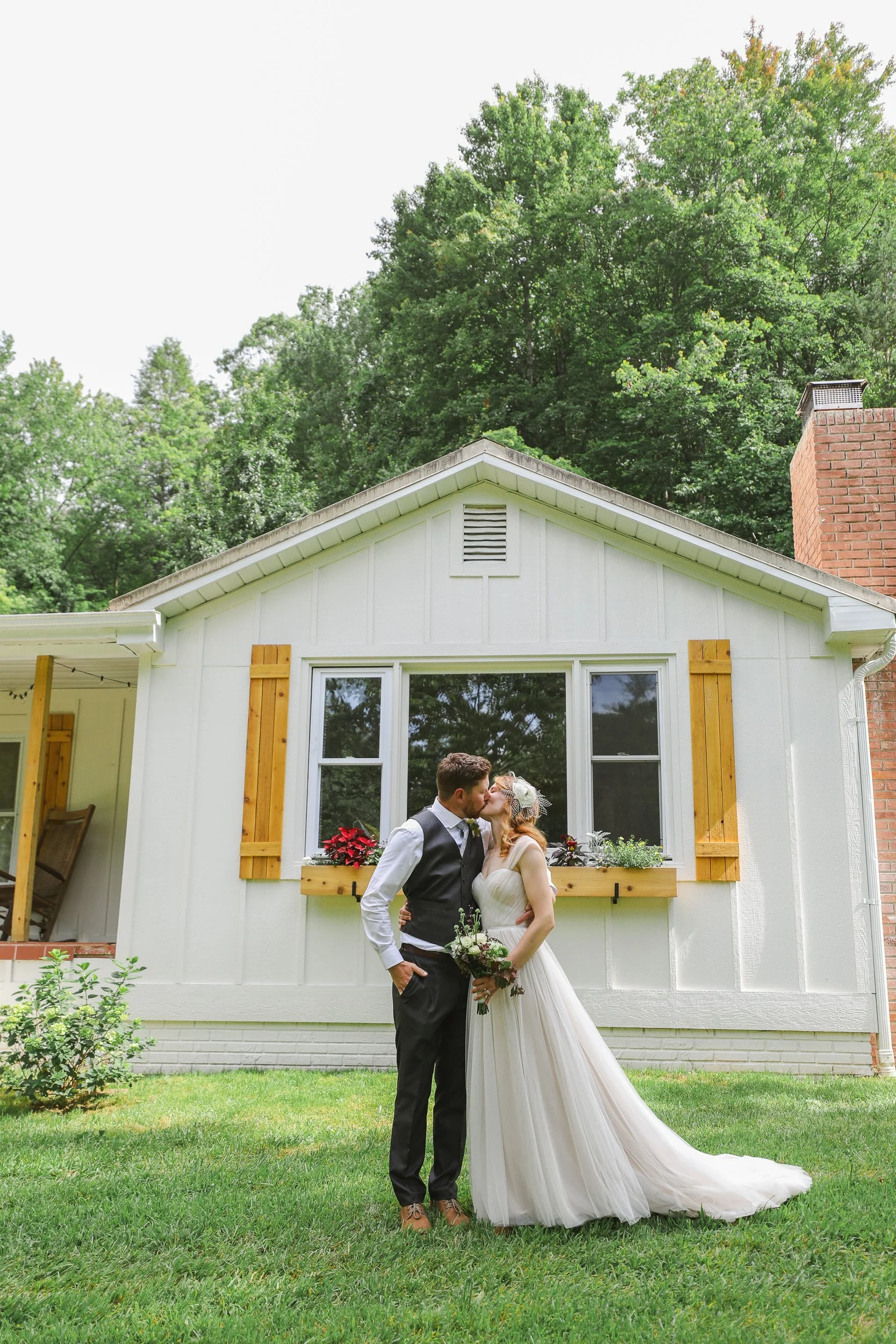 A couple in wedding attire standing outdoors in front of a white house, sharing a kiss with greenery and trees in the background in Greenville, SC