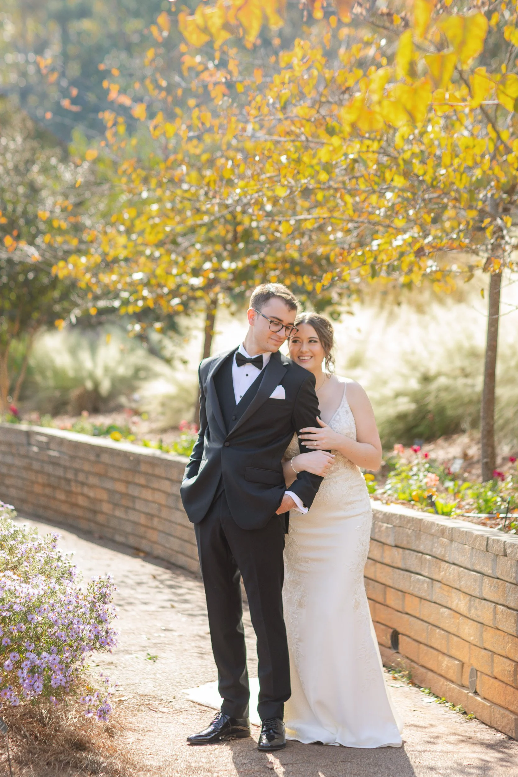 A newlywed couple in wedding attire standing outdoors with colorful autumn trees in the background. The groom is wearing a black tuxedo with a bow tie, and the bride is in a white lace wedding dress, smiling and holding onto the groom's arm.