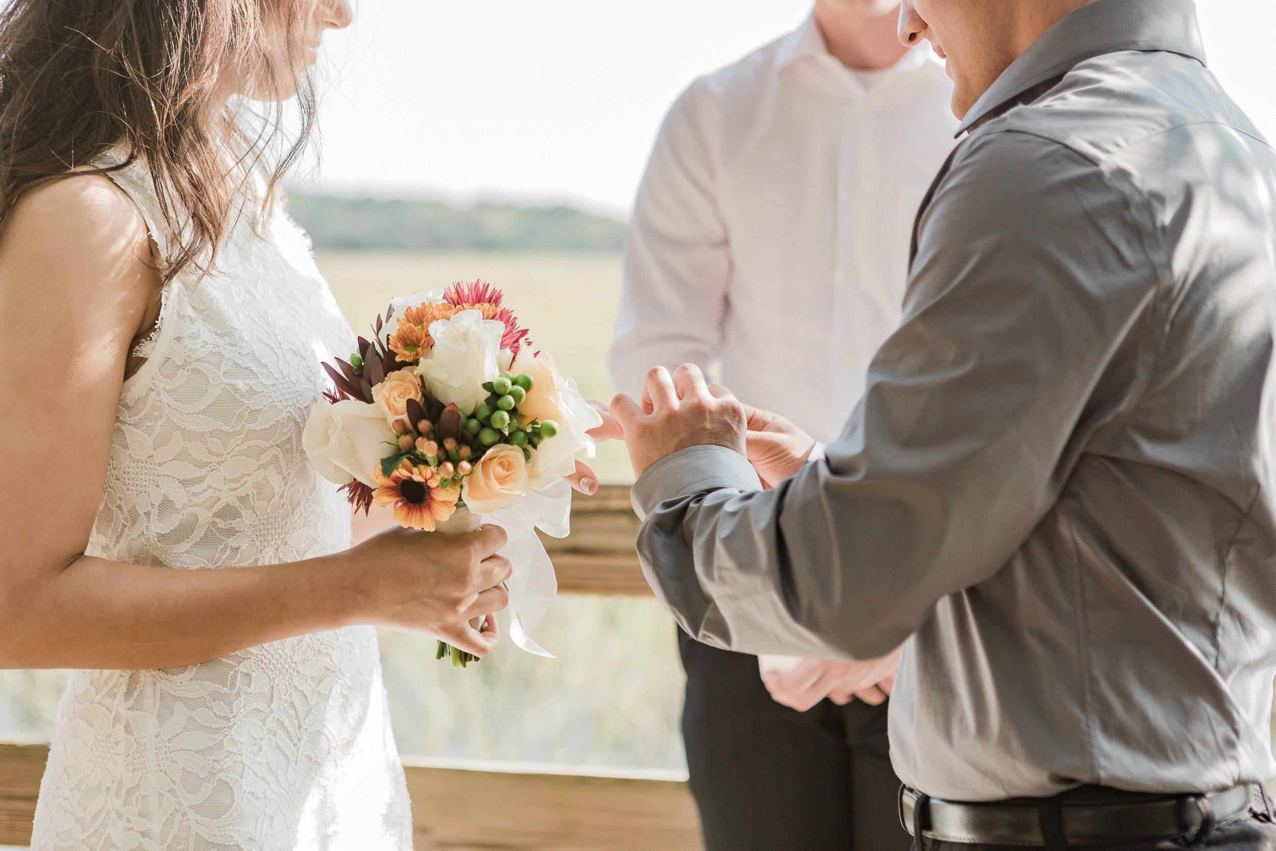 A couple is exchanging wedding rings during a ceremony, with an officiant present. The bride is holding a bouquet of flowers, and the groom is wearing a gray shirt outdoors during elopement in Greenville, SC