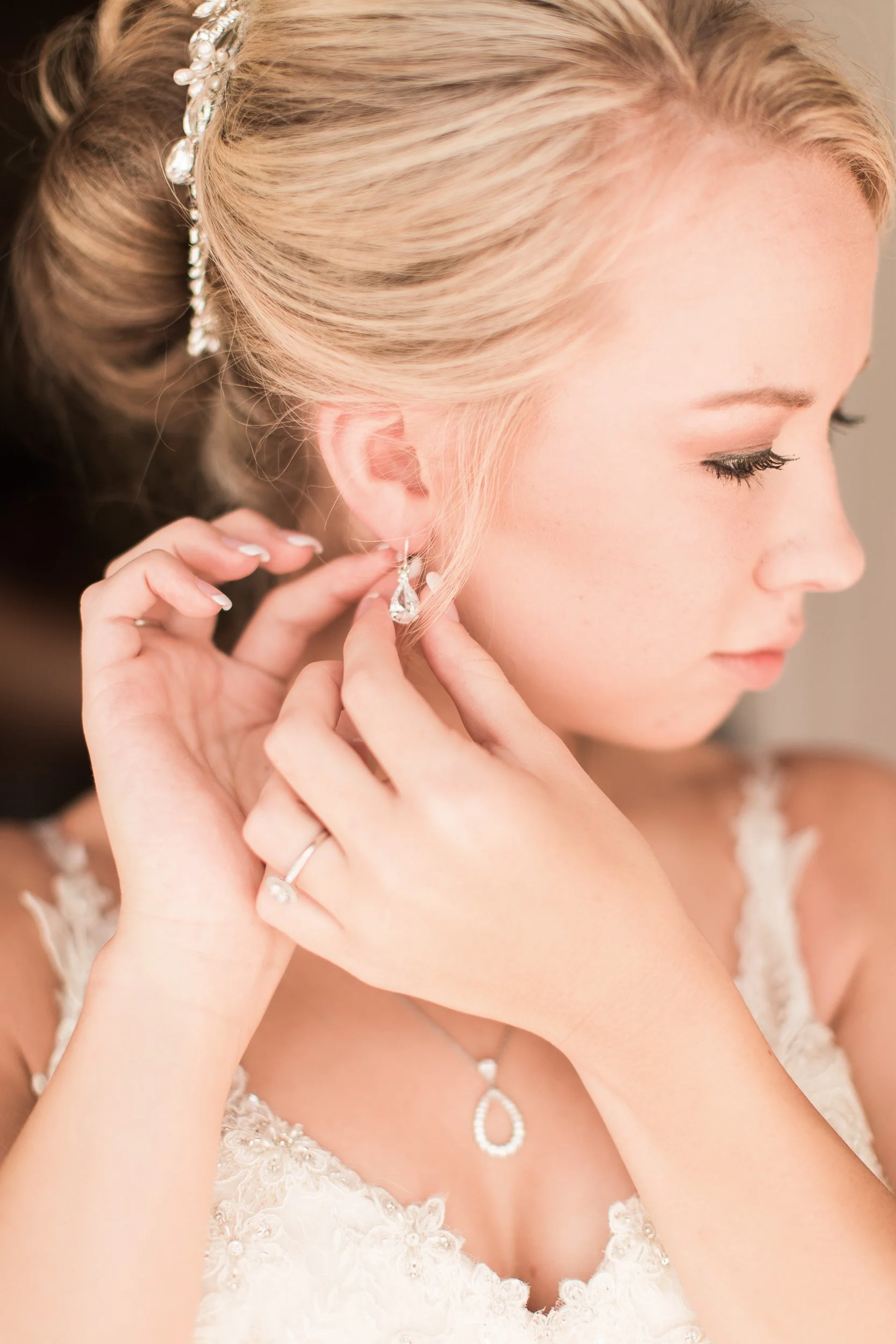 A woman with blonde hair styled in an updo, wearing a decorative hairpiece, and getting ready for a special occasion by putting on earrings before intimate wedding at Lake Keowee in South Carolina