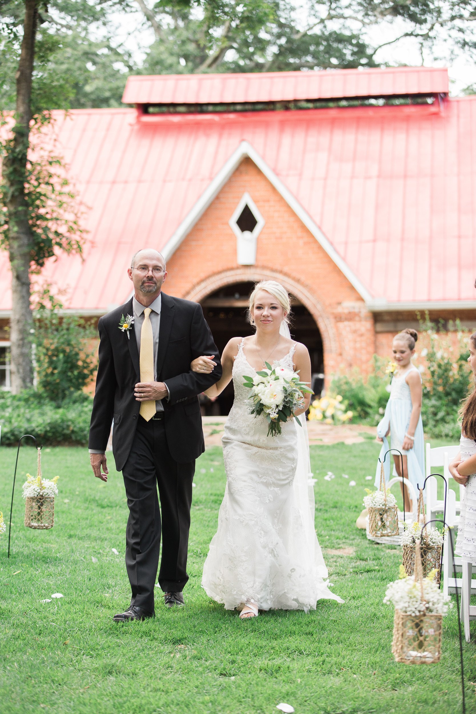 A bride in a white wedding dress holding a bouquet walks down the aisle assisted by father during intimate wedding at Sleepy Hollow Barn in Pendleton, SC