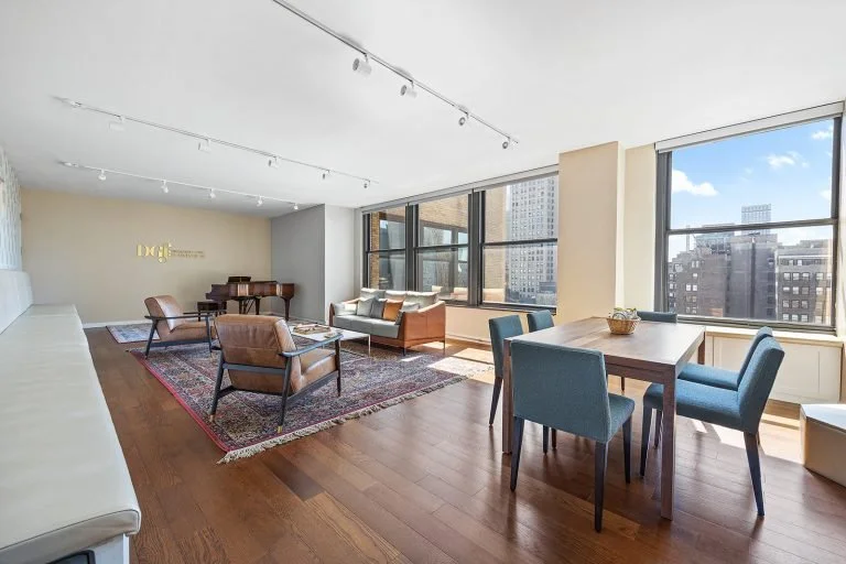 This image depicts a modern, well-lit rehearsal space with large windows offering a city view, furnished with a mix of comfortable seating, a dining table with chairs, and a piano in the corner, set on hardwood floors with a vibrant area rug.