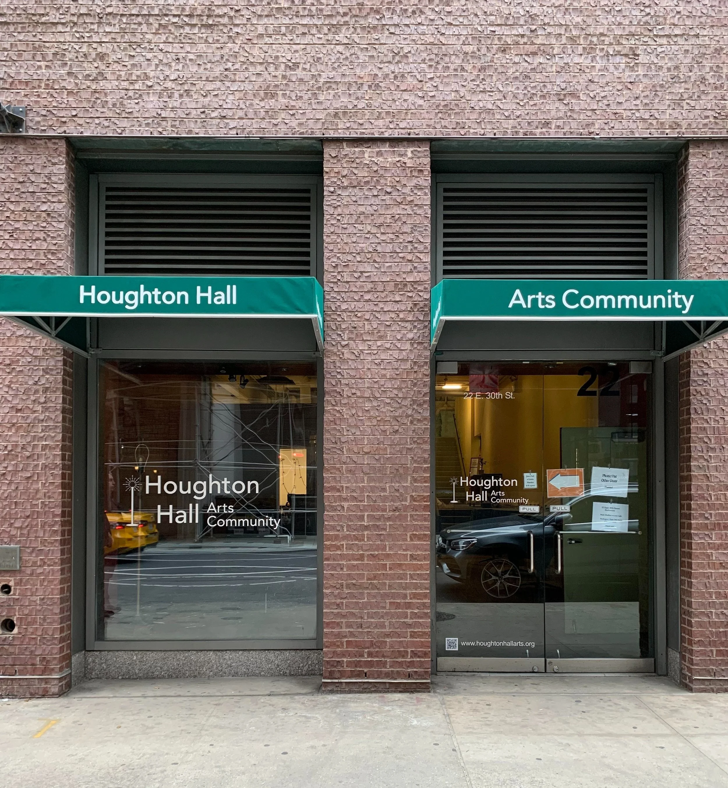 Photograph of the entrance to Houghton Hall, part of the Arts Community. The building facade is brick, with two glass doors under green awnings; one reads 'Houghton Hall' and the other 'Arts Community.' A reflection of the street and parked cars can