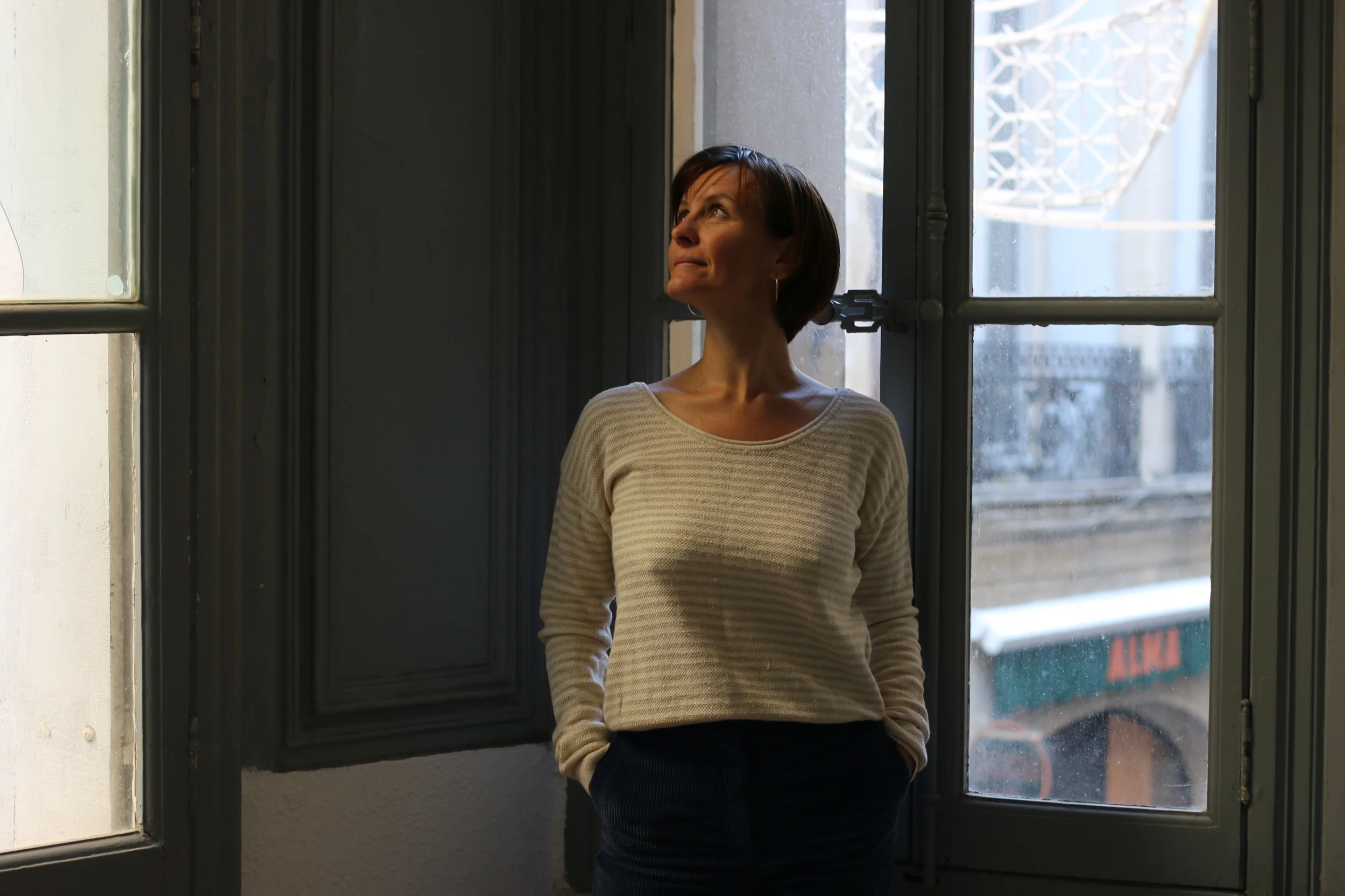 Femme debout près d'une fenêtre, regardant vers le haut, dans une pièce avec lumière naturelle.