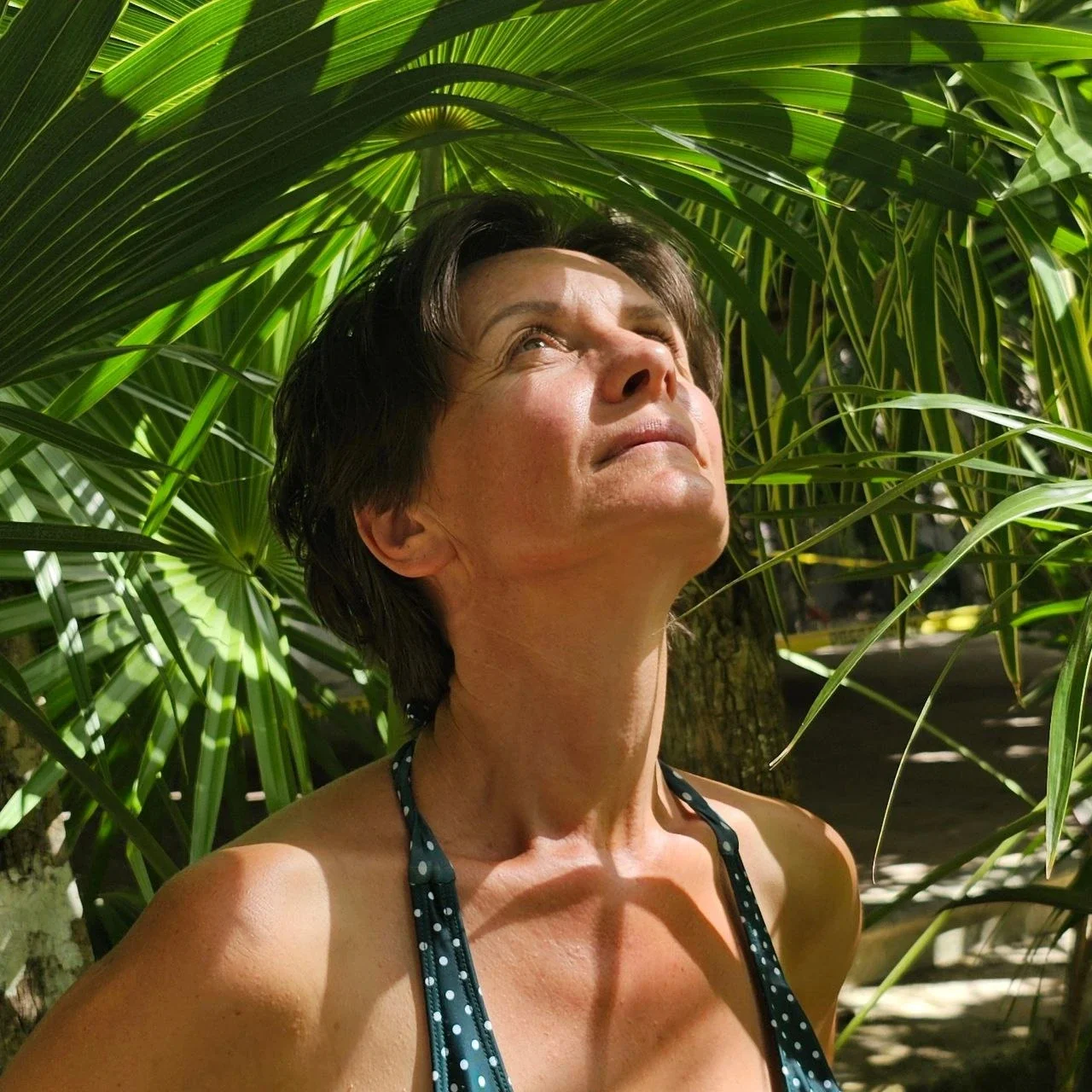 Femme debout sous le soleil parmi des palmiers, regardant vers le haut avec un visage serein.