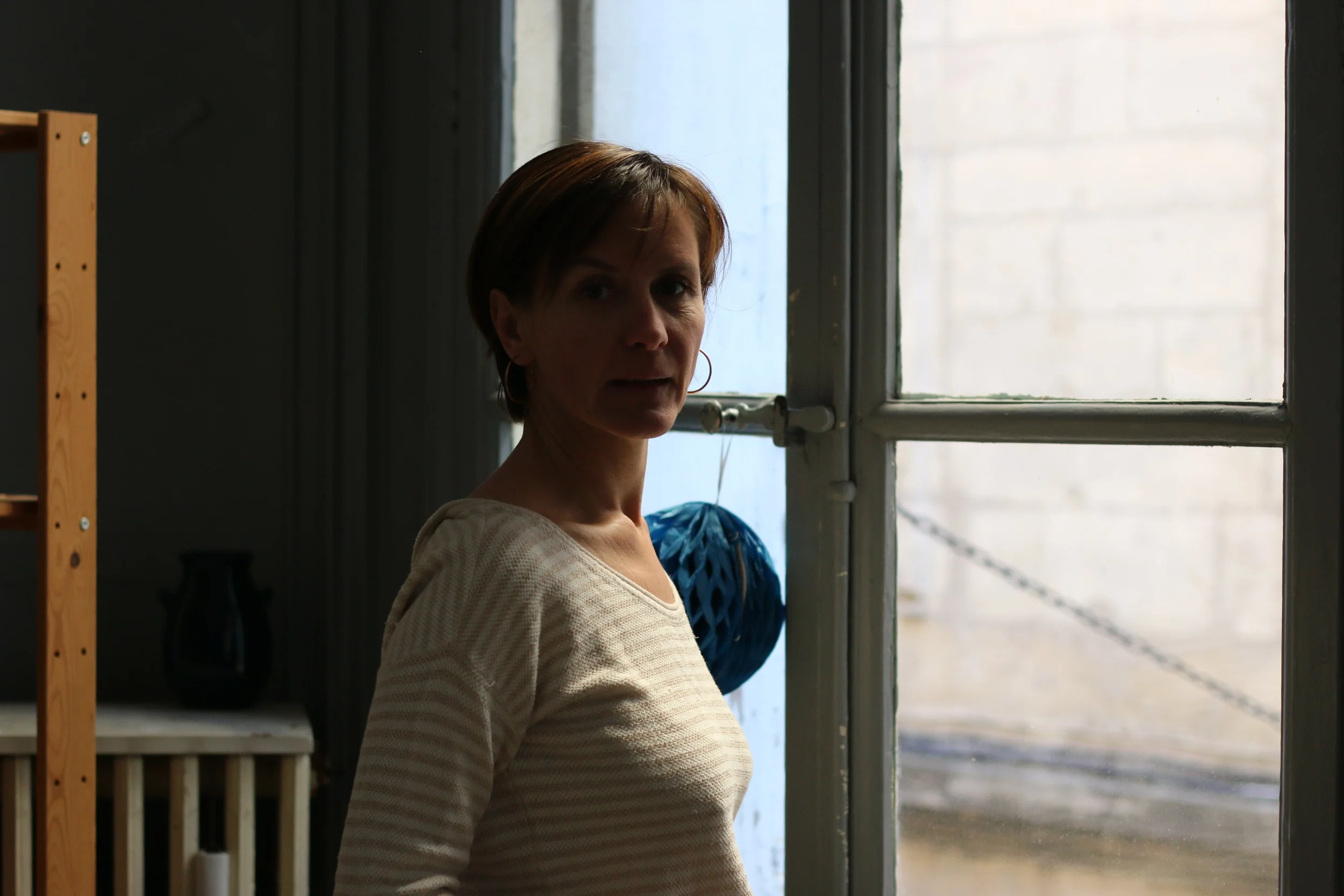 Une femme avec des cheveux courts bruns porte un pull beige à rayures, se tient à côté d'une fenêtre dans une pièce sombre, avec une vue partielle d'une pièce en arrière-plan.