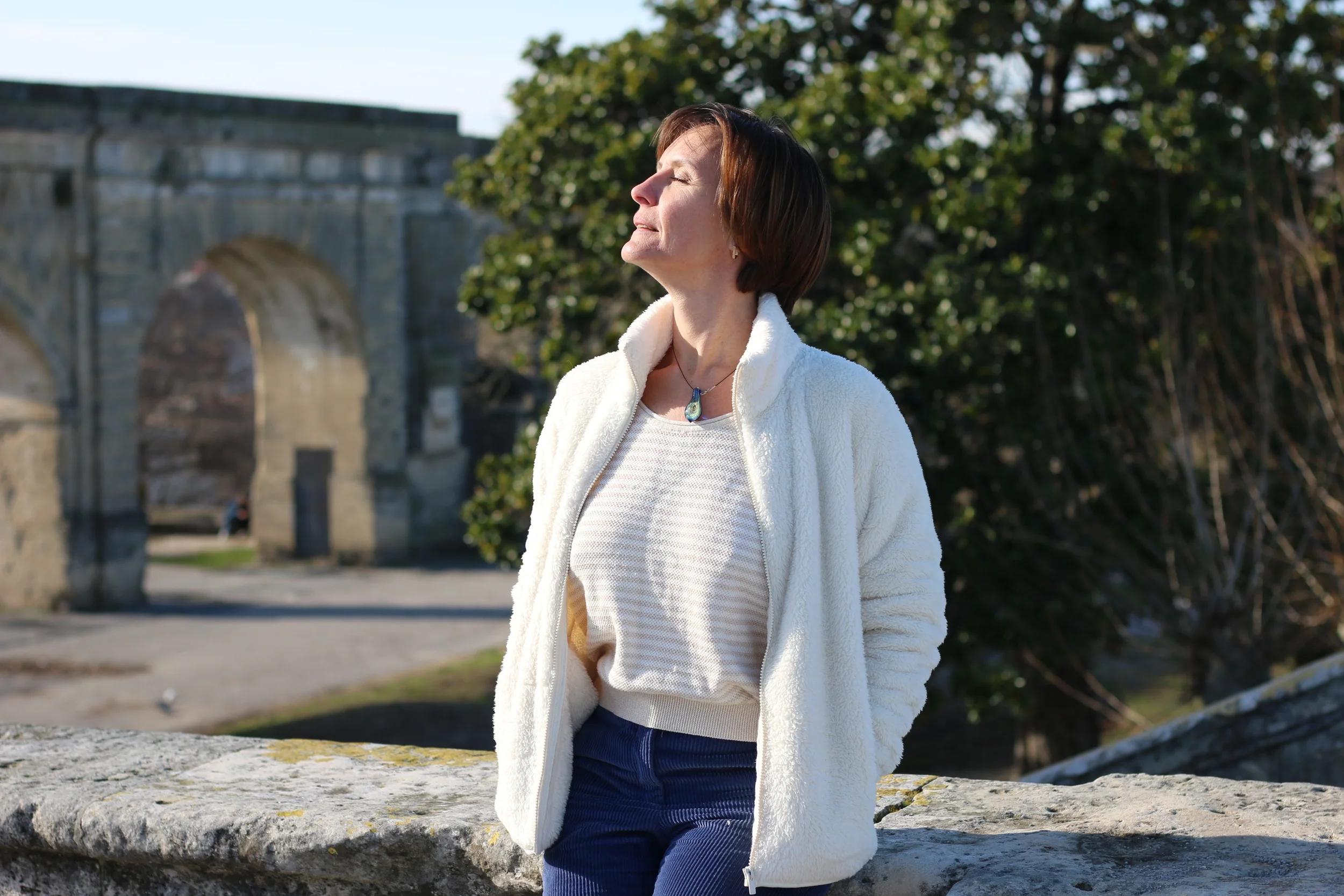 Une femme debout en plein air, profitant du soleil avec les yeux fermés, portant un pull blanc et un collier, devant une structure ancienne en pierre et des arbres.