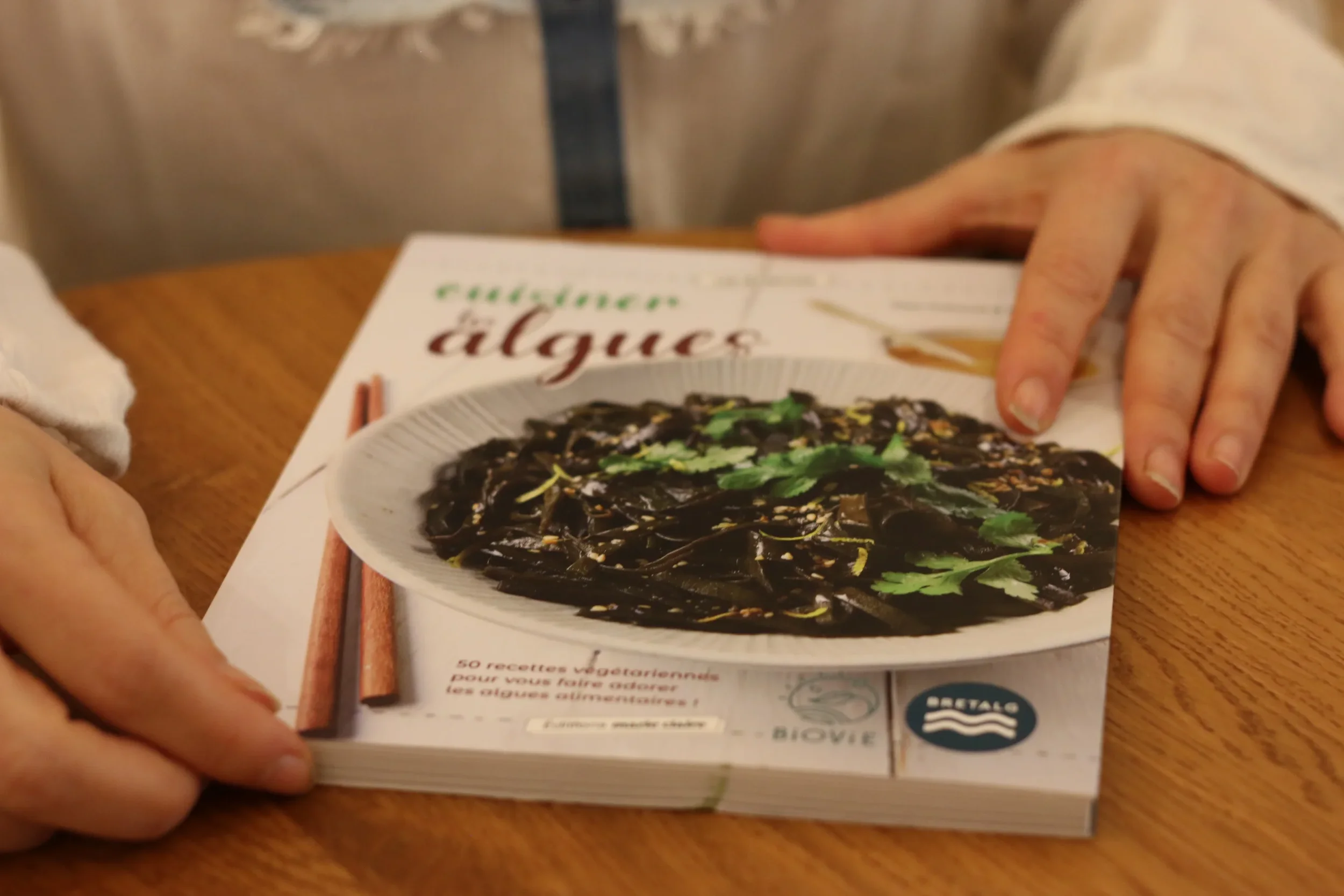 Une personne tient un livre de cuisine ouvert montrant une recette végétarienne avec des nouilles sombres et des herbes, sur une table en bois.