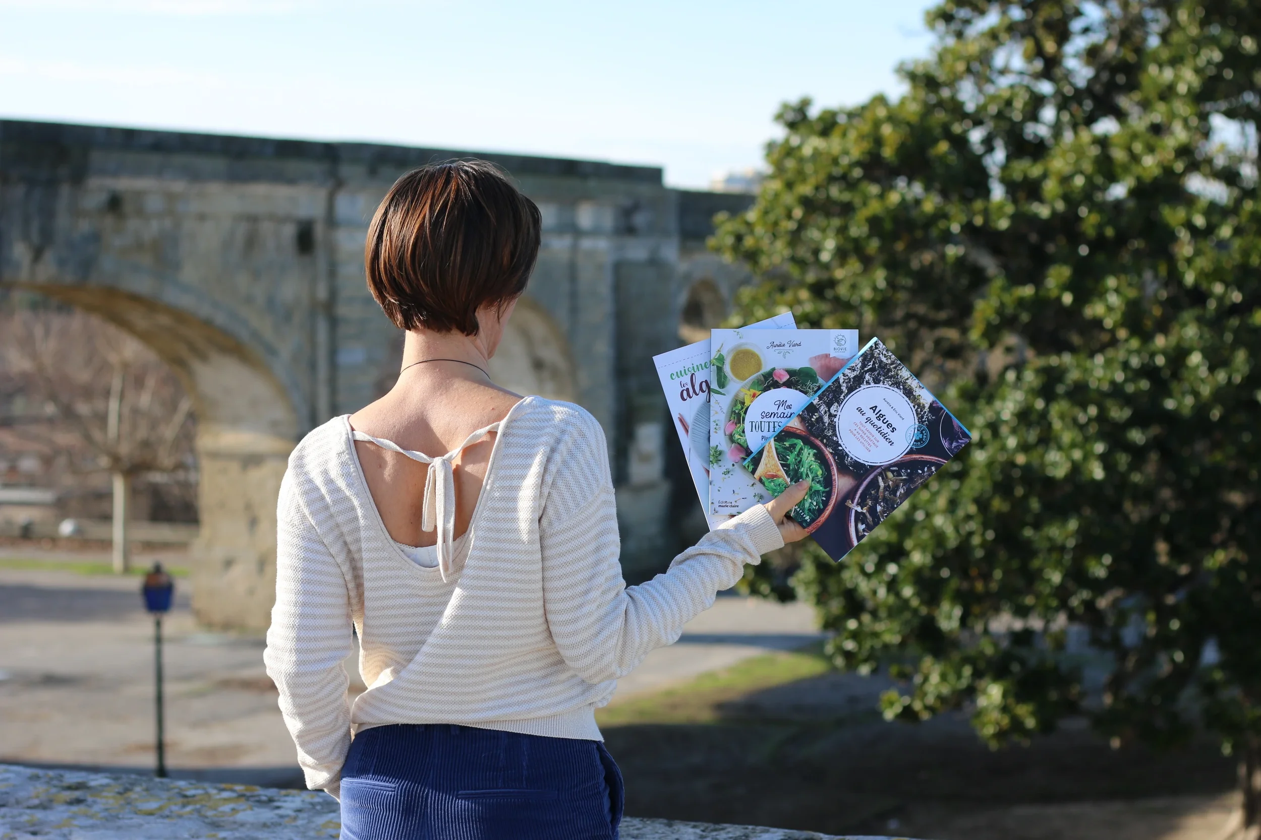 Une femme vue de dos, tenant des brochures ou magazines dans une main, en extérieur devant un pont en pierre et des arbres.