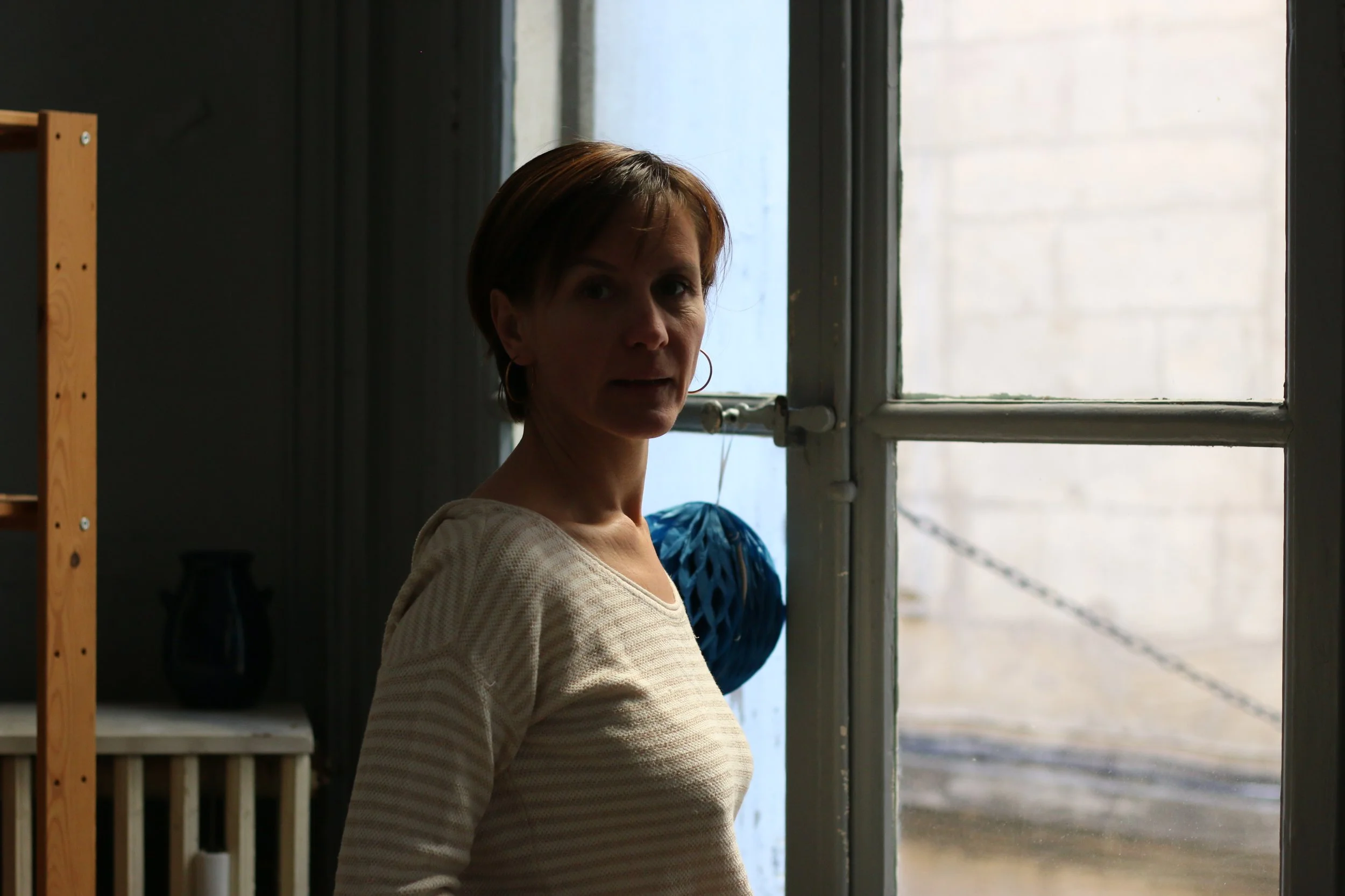 Une femme avec des cheveux courts et bruns, portant un haut crème, se tient devant une fenêtre dans une pièce sombre. Il y a une étagère en bois avec des objets noirs à gauche.