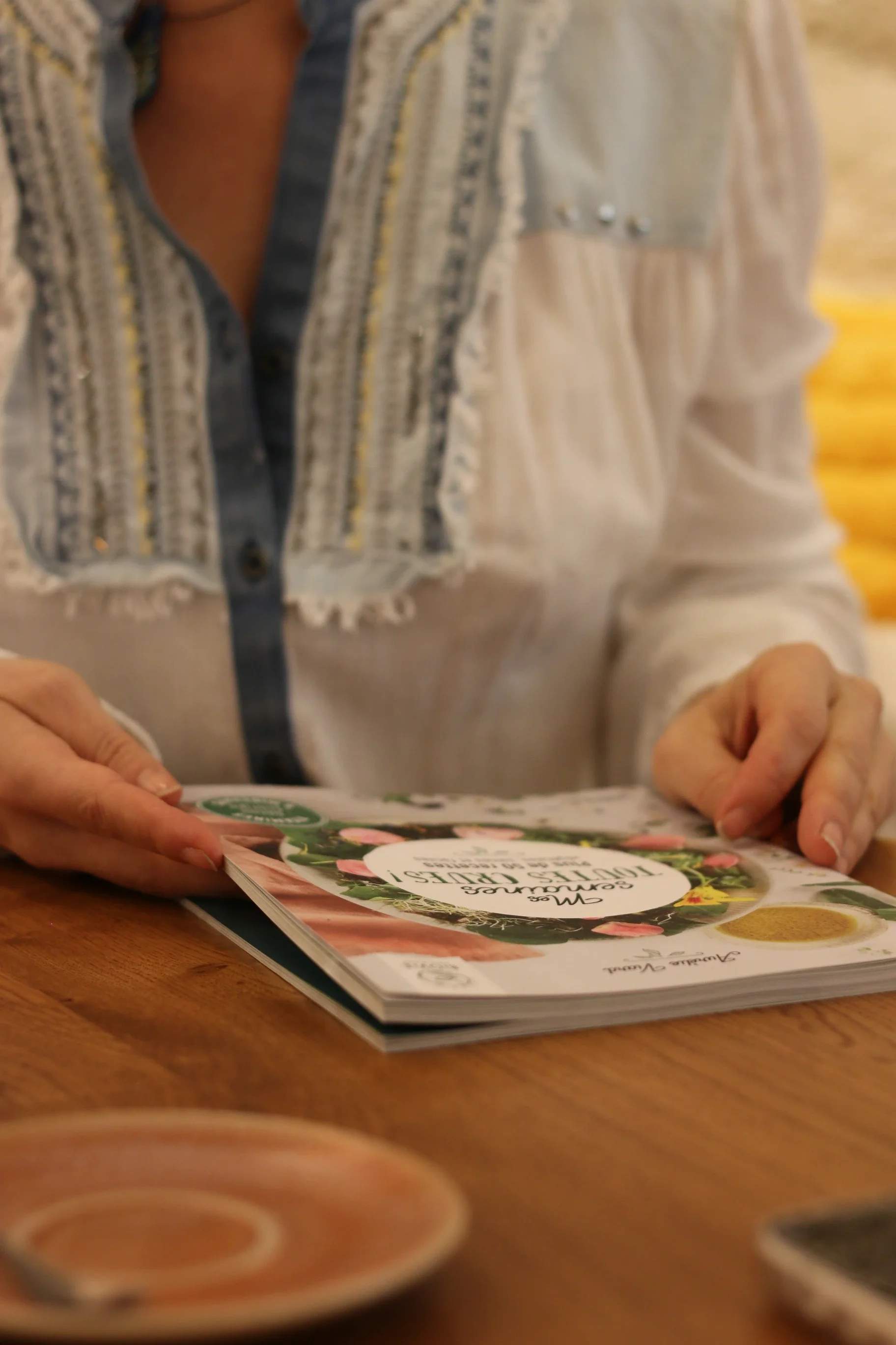 Une personne tient un magazine de cuisine sur une table en bois, le magazine est ouvert avec des images de plats et des textes.