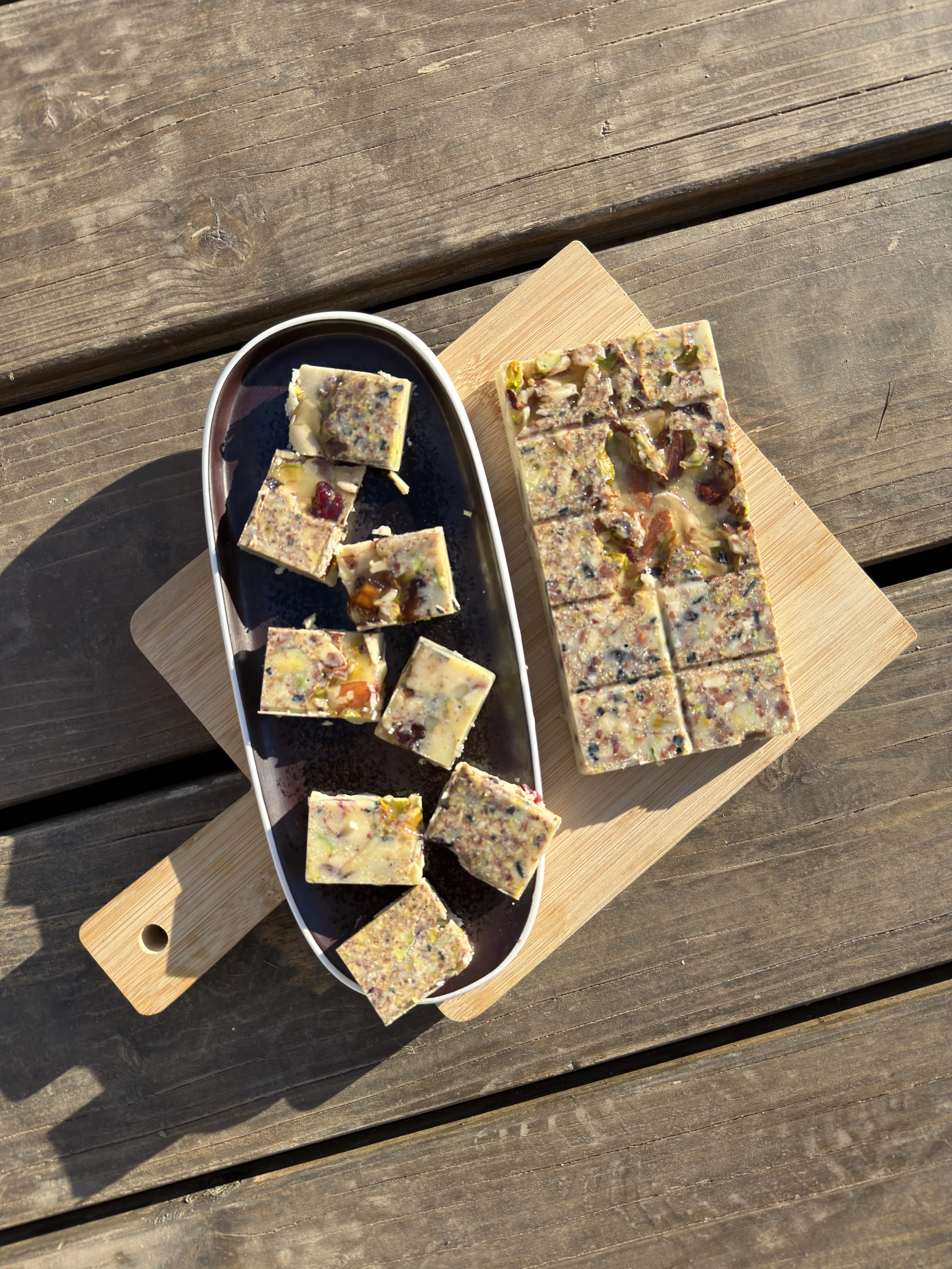 Tablette de chocolat blanc aux noix et fruits séchés, avec quelques morceaux coupés et placés sur une petite assiette, sur une planche en bois.