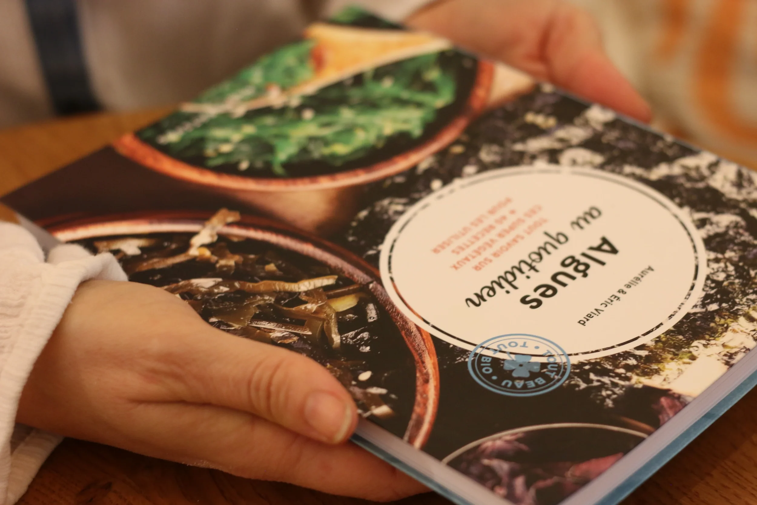 Une personne tient un livre de cuisine végétarienne sur un fond de table en bois. La couverture montre des plats végétaux et possède un label bio.