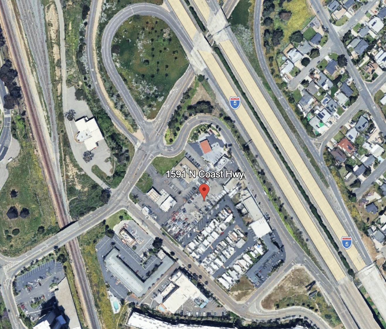 Satellite view of a city area showing roads, buildings, a parking lot, a roundabout, a park, and nearby transportation infrastructure including railway tracks and highways.