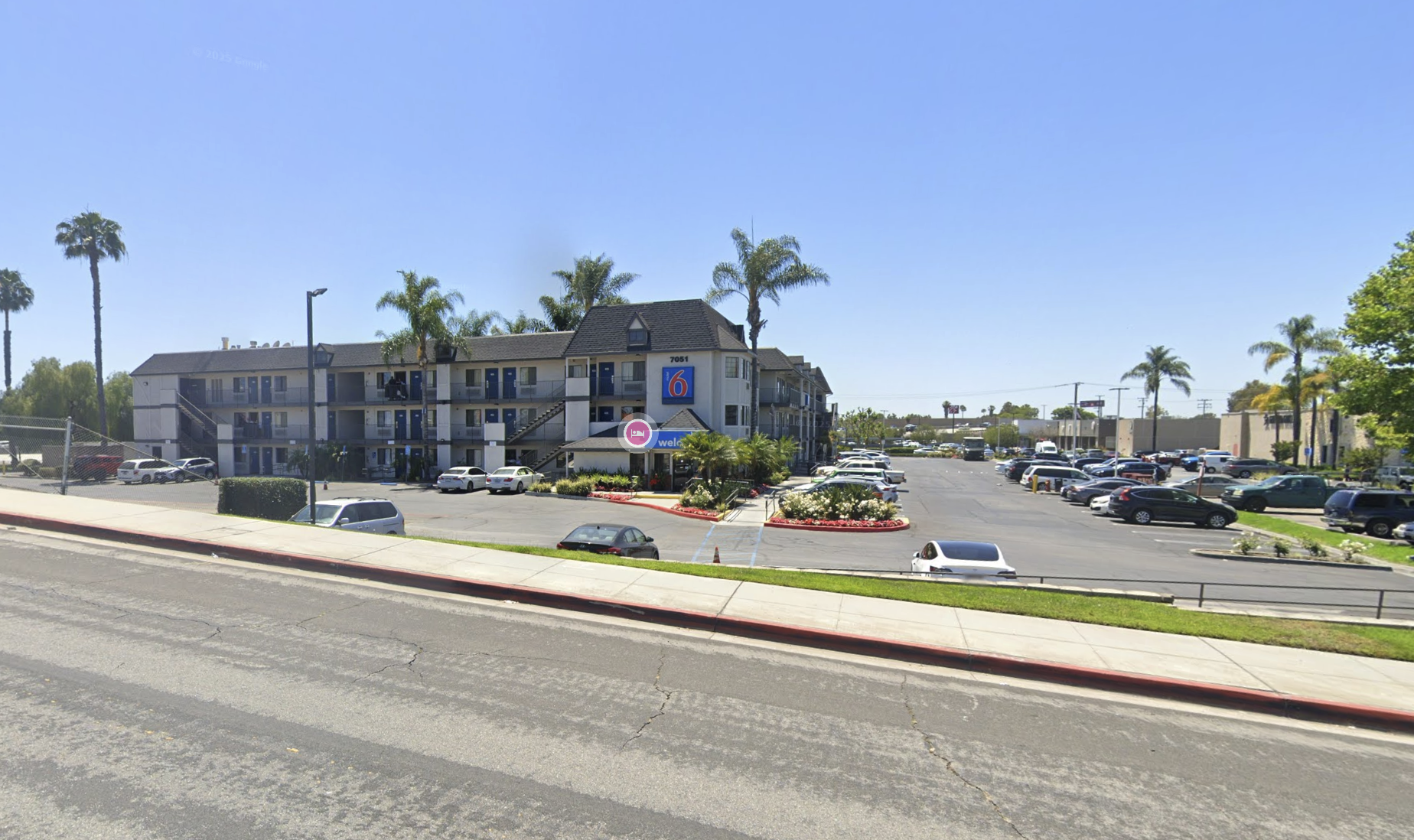 Parking lot in front of a multi-story motel with palm trees, blooming bushes, and a large "6" sign on the building, under a clear blue sky.