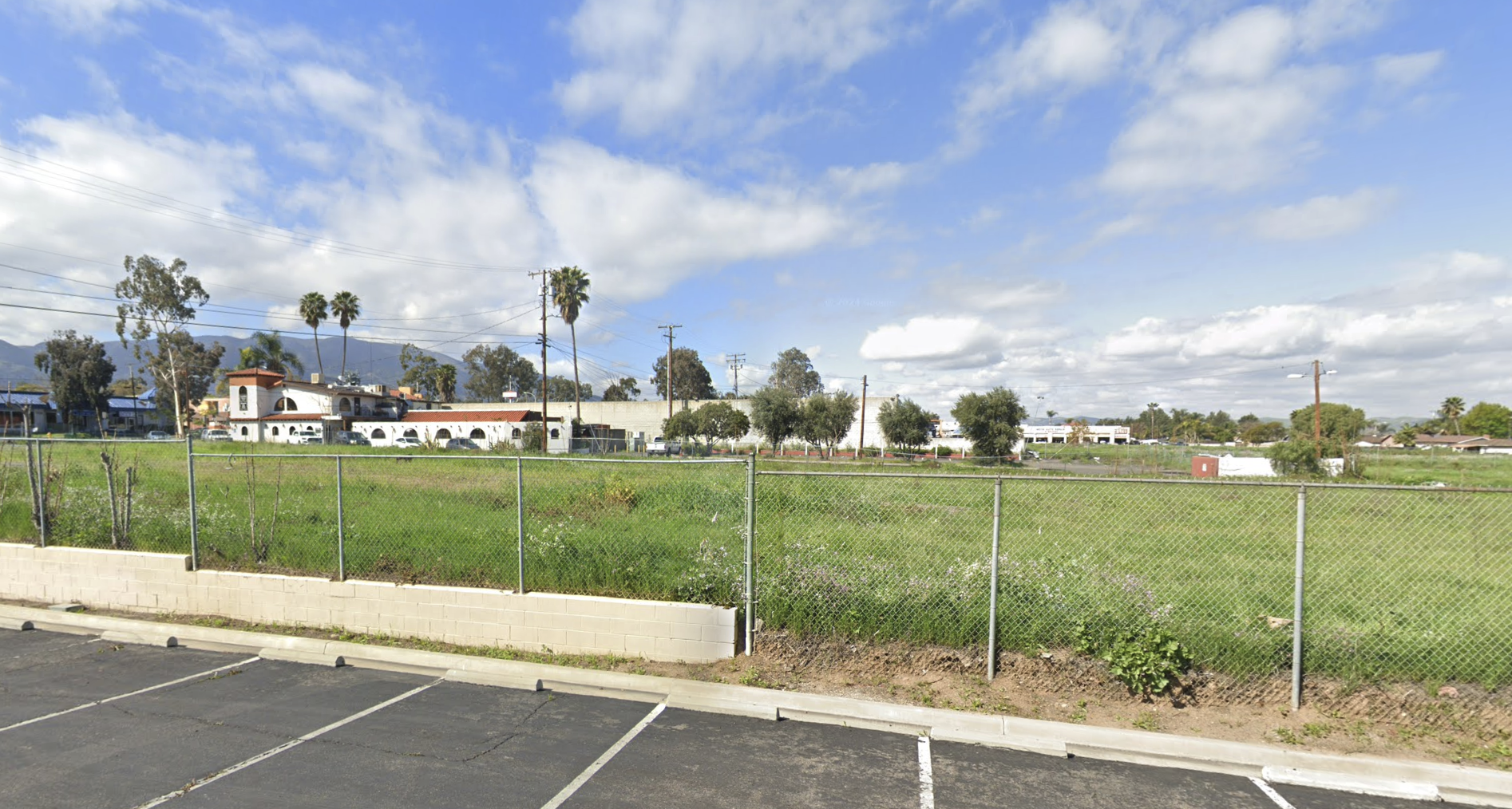 A parking lot in the foreground with empty spaces, a chain-link fence behind it, green grass, and trees in the background under a partly cloudy sky.