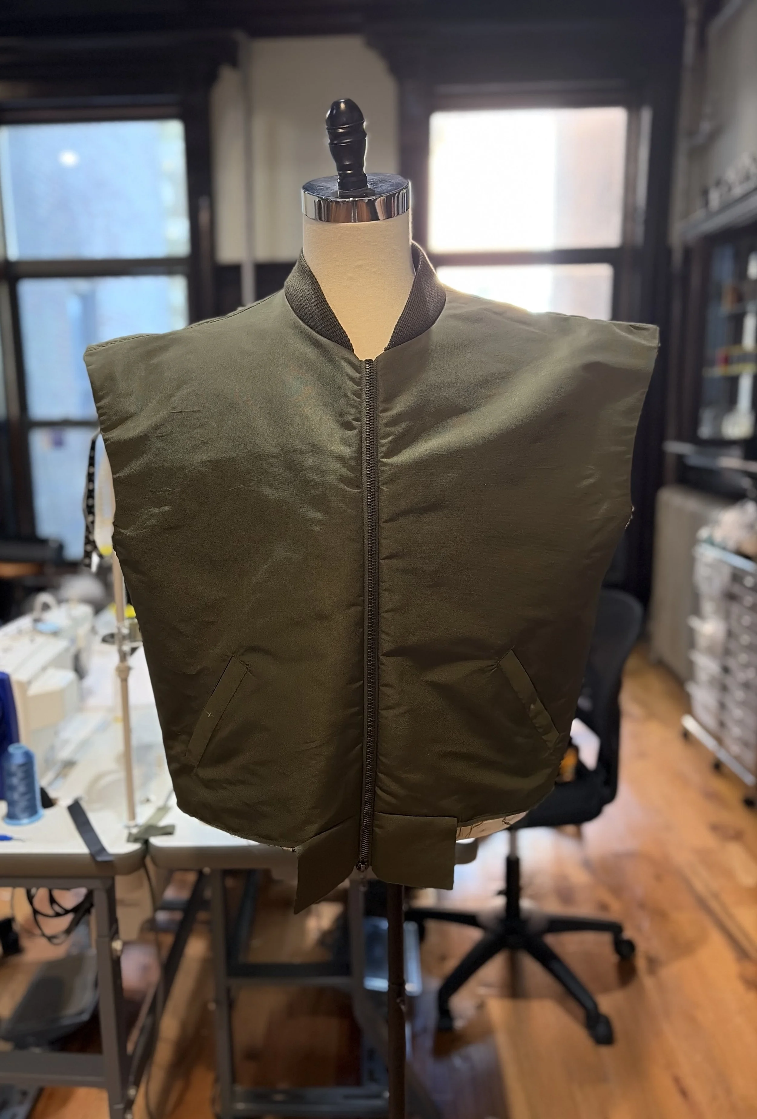 A mannequin torso wearing a green bomber jacket with black accents on the collar, displayed on a stand in a workshop with windows in the background.
