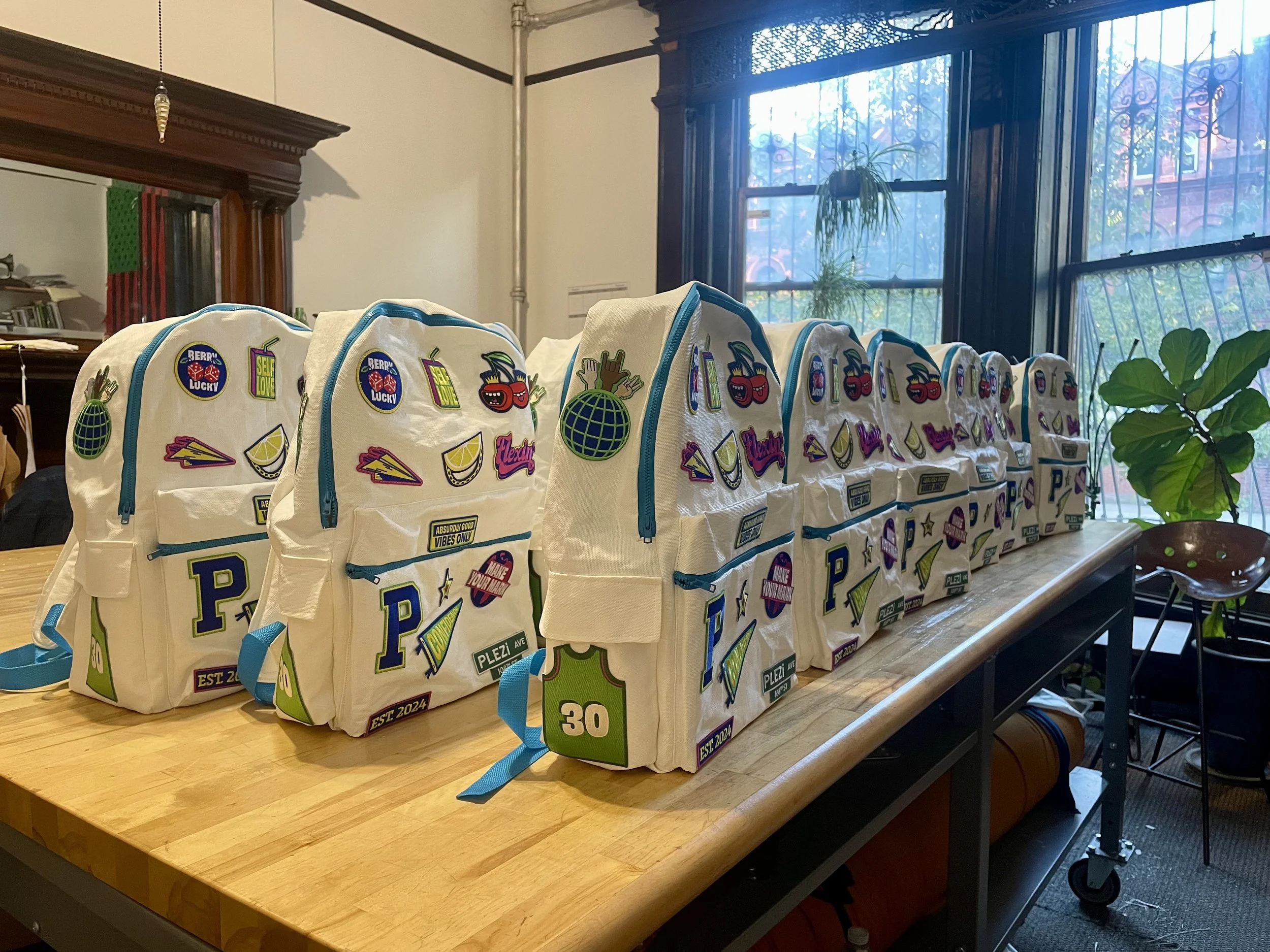 Multiple white backpacks with colorful patches and zippers are lined up on a wooden table, near a window with plants outside and a large mirror in the background.