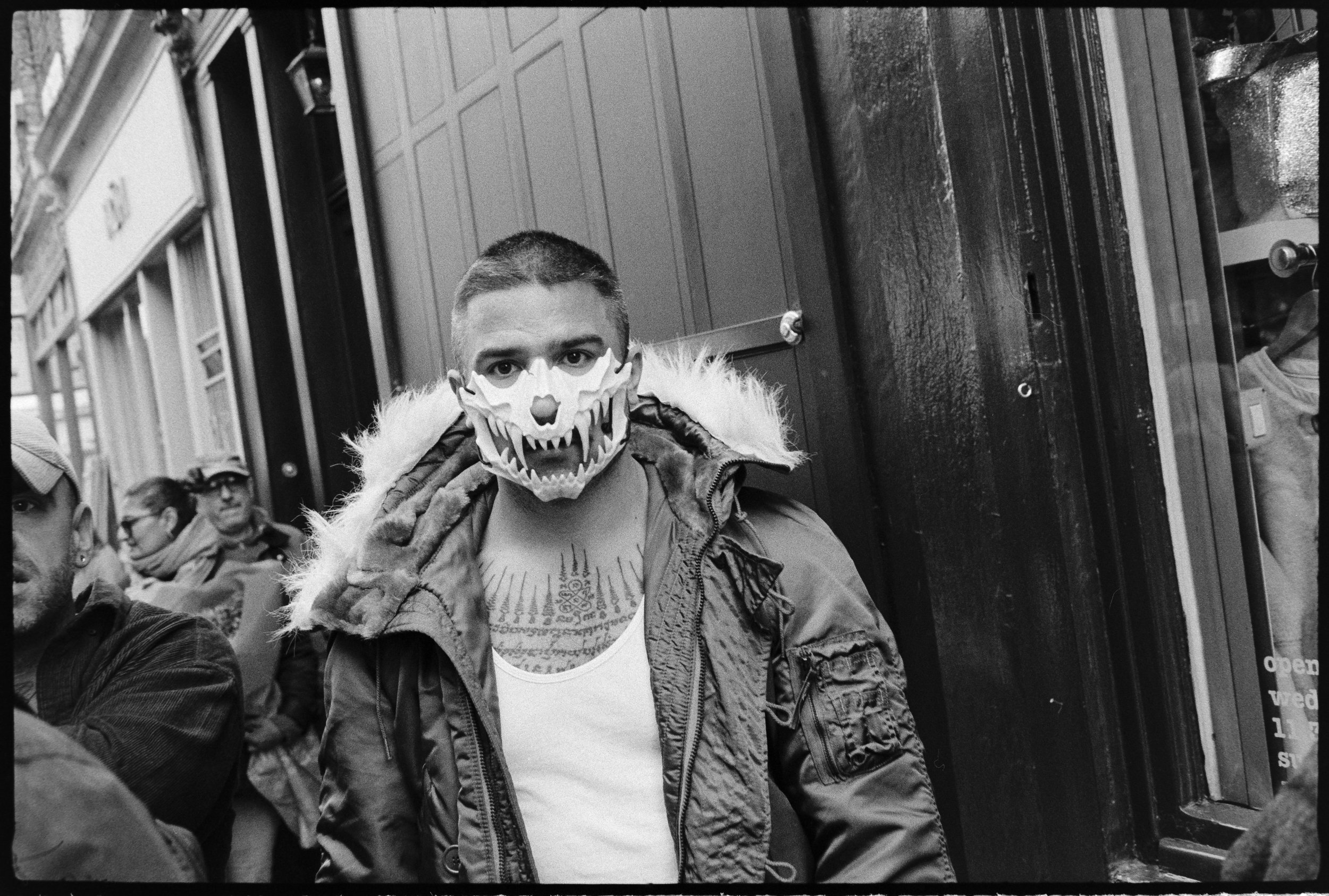 A man wearing a white tank top and a jacket with a fur-lined hood stands on a city street, wearing a skull mask with sharp teeth and a hollow nose. There are other people in the background, including a woman with glasses and a scarf.