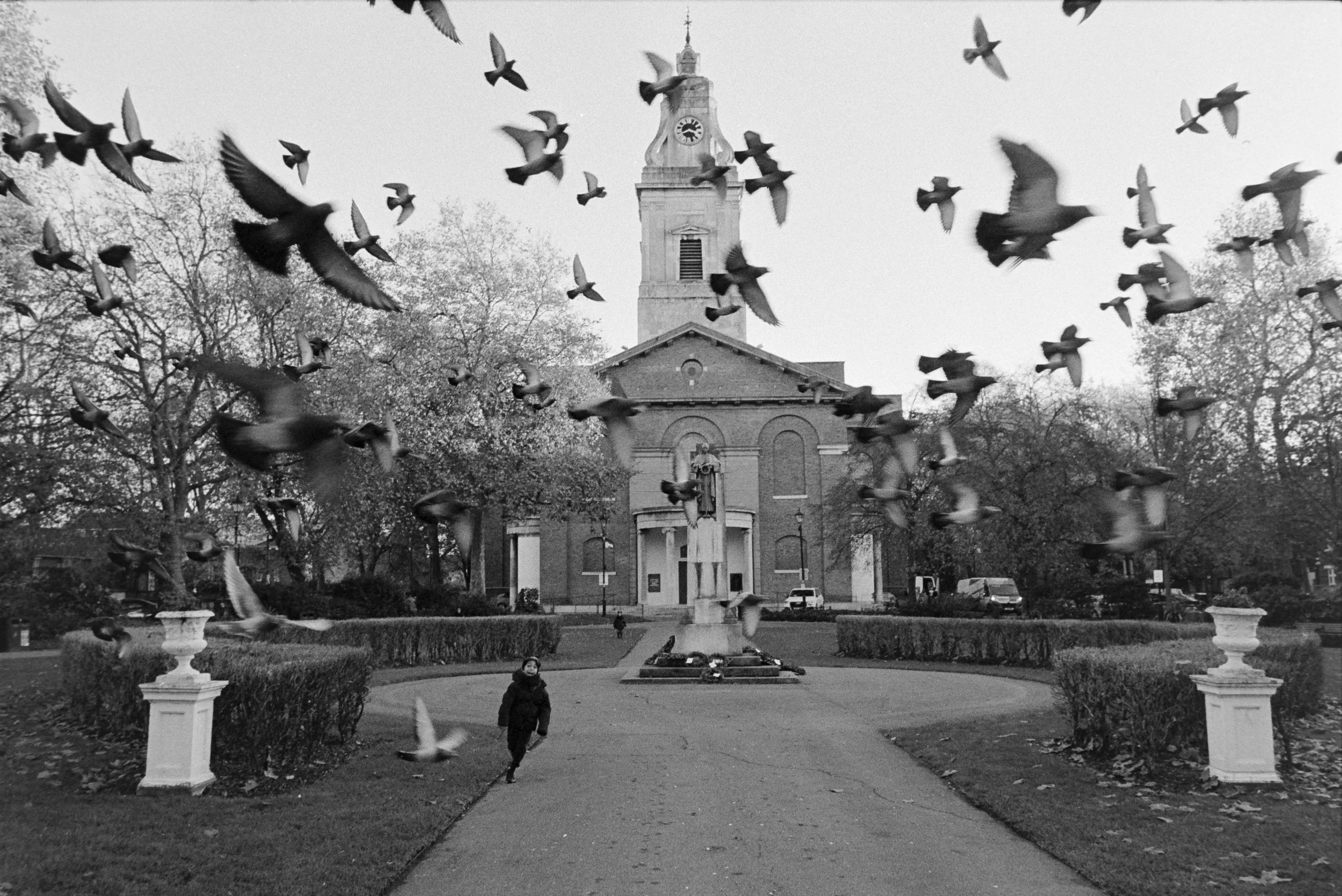 Black and white photo of a church with a tall clock tower, a statue in front, and a child running along a paved path, with many pigeons flying overhead.