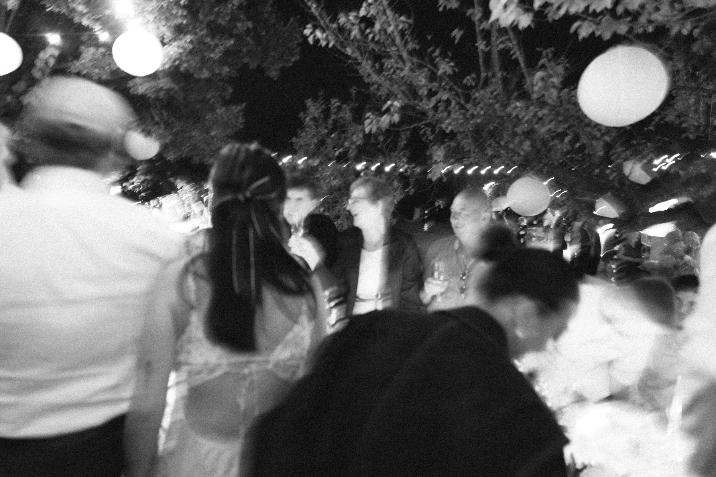 Group of people socializing at night under decorative hanging lanterns with trees in the background.
