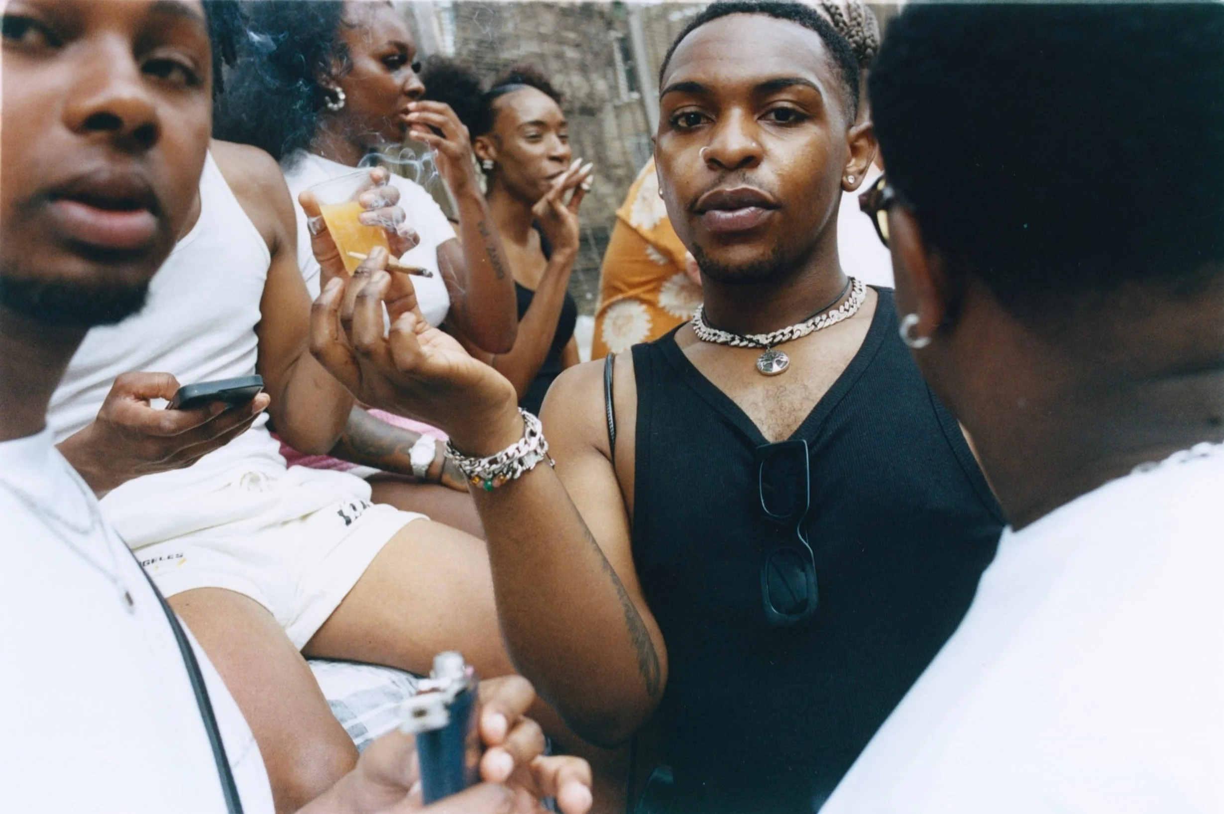 Group of people socializing and smoking outdoors, including a woman in a black top with jewelry, and others holding drinks and cigarettes.