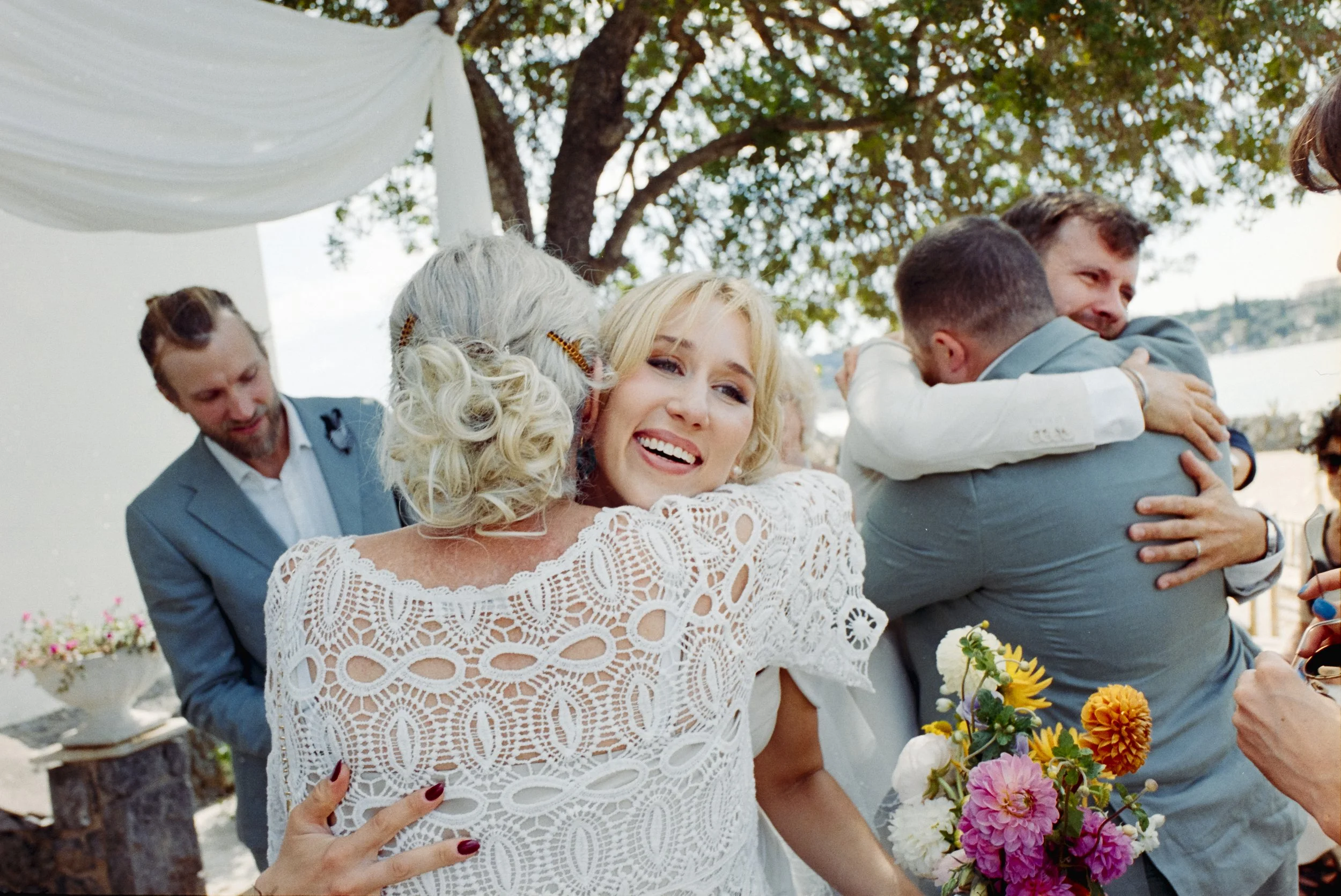 Group of people at an outdoor wedding, hugging and smiling, with a woman holding a colorful bouquet of flowers, surrounded by greenery and a view of water in the background.