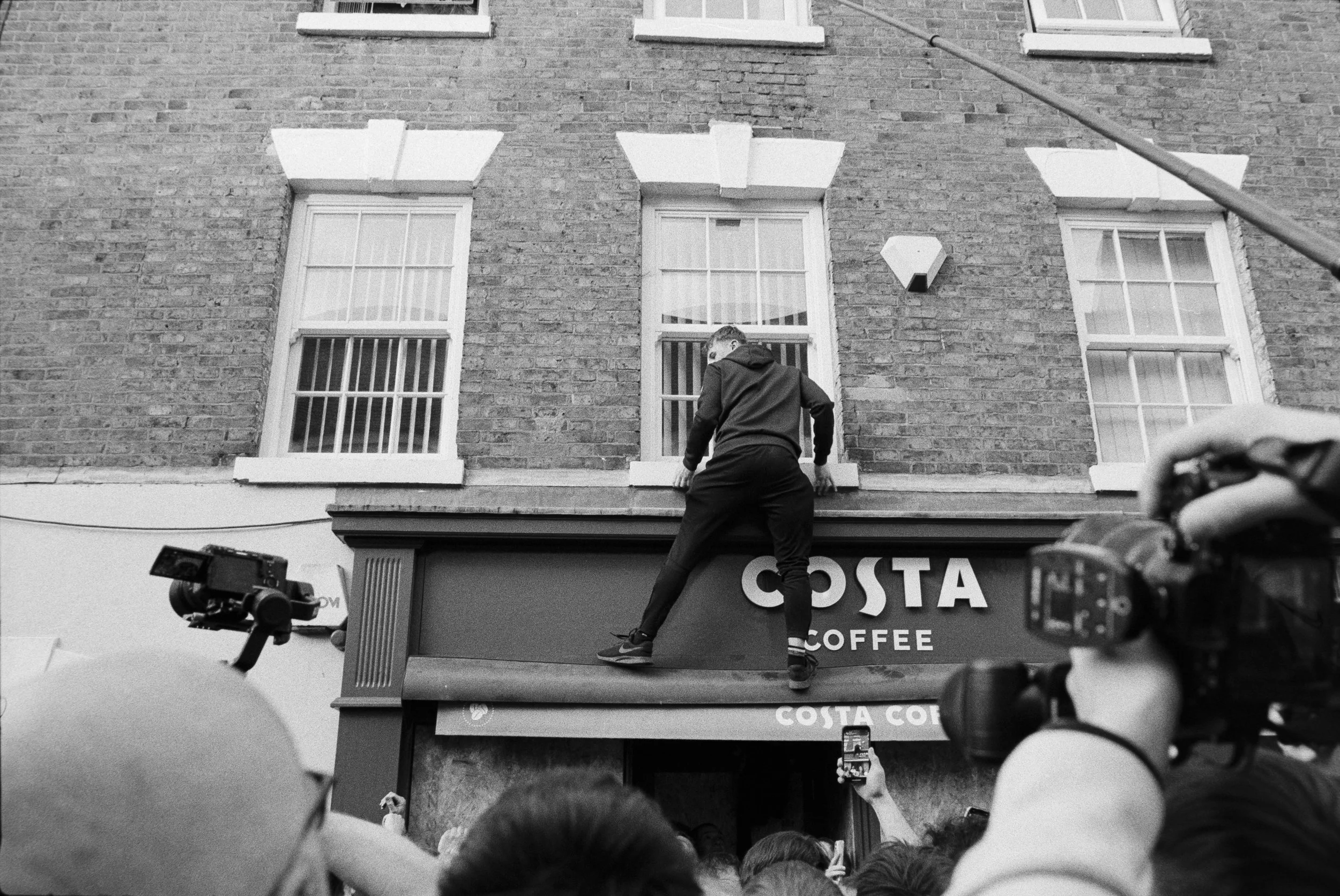 A young man climbing on the sign of a Costa Coffee storefront in an urban area while a crowd watches and takes photos. The building has three windows and a brick exterior.