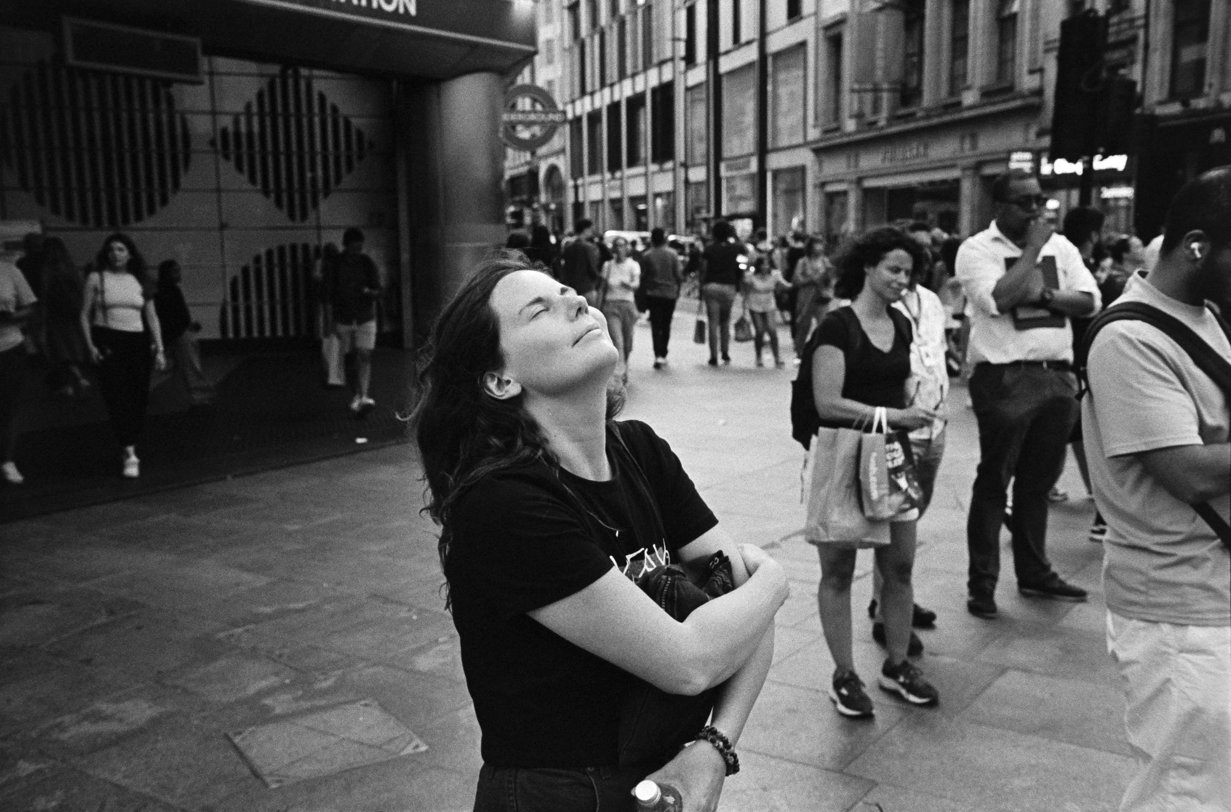 A woman with eyes closed and arms crossed stands in a crowded city street, surrounded by people and tall buildings, in a black and white photo.