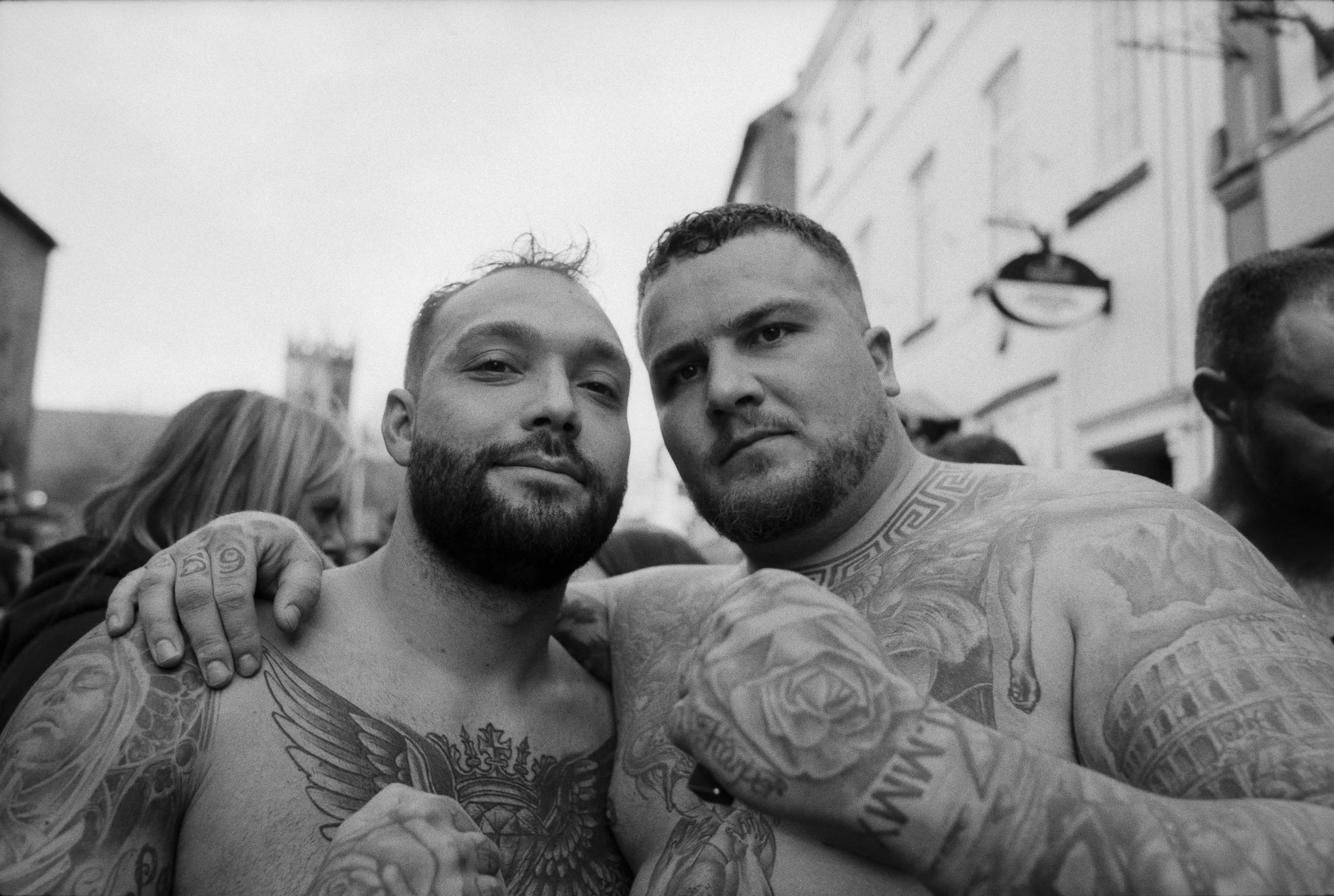 Two tattooed shirtless men with beards taking a selfie together outdoors in a crowd, with buildings in the background.