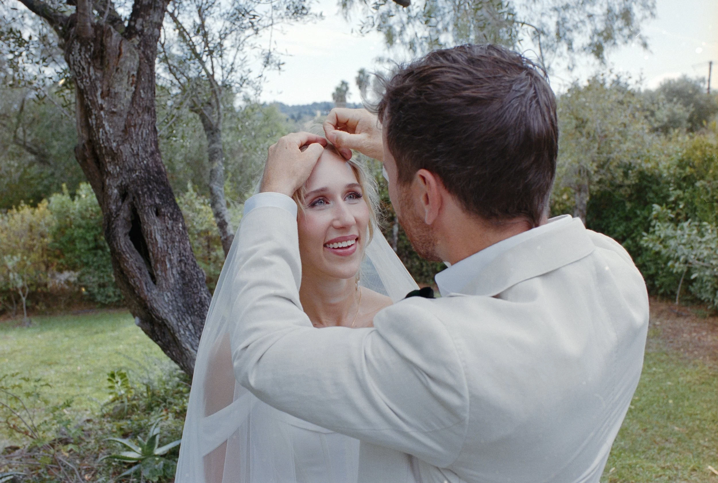A man is placing a wedding ring on a woman's finger outdoors, surrounded by trees and greenery, during a wedding ceremony.