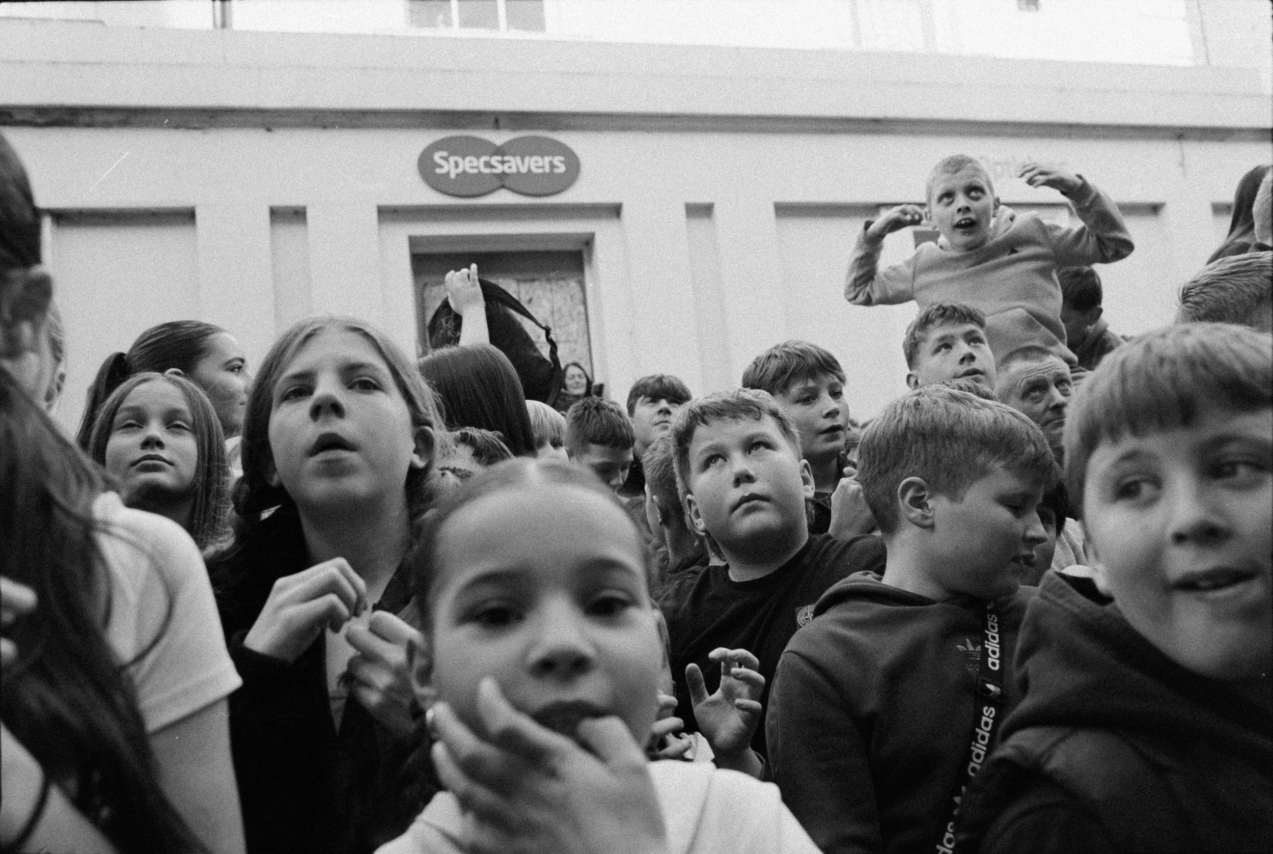 A crowd of children and a few adults seated in an indoor setting, some looking upwards and some with curious expressions, with a "Specsavers" sign visible in the background.