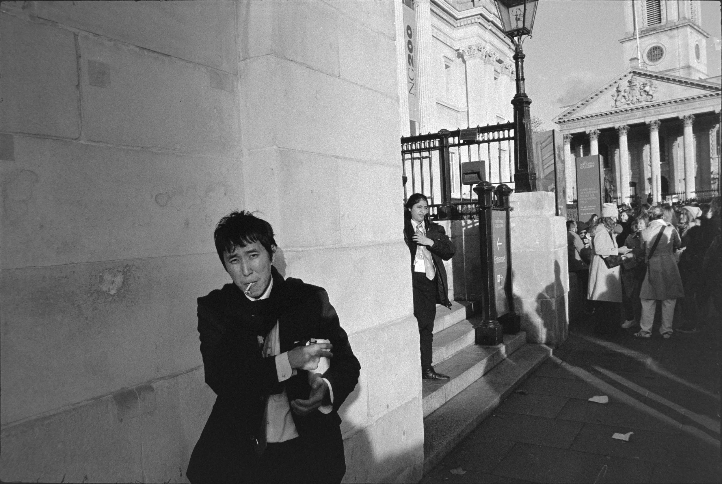A black and white photo of a man with a cigarette in his mouth and a woman standing on steps near a historic building with columns and a crowd of people in the background.