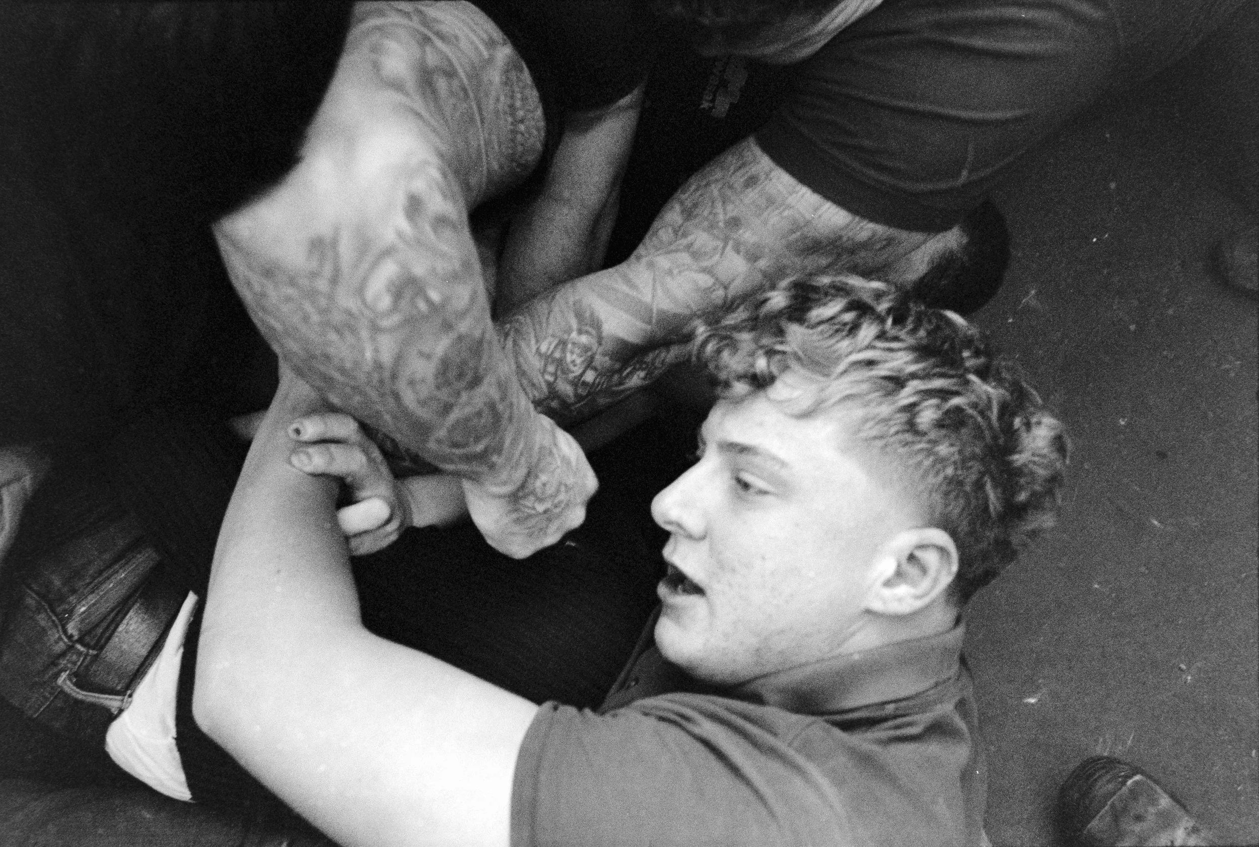 A young man with curly hair and tattoos on his arms is lying on the ground, engaging in a physical altercation with another person. The young man appears to be trying to defend himself or break free.