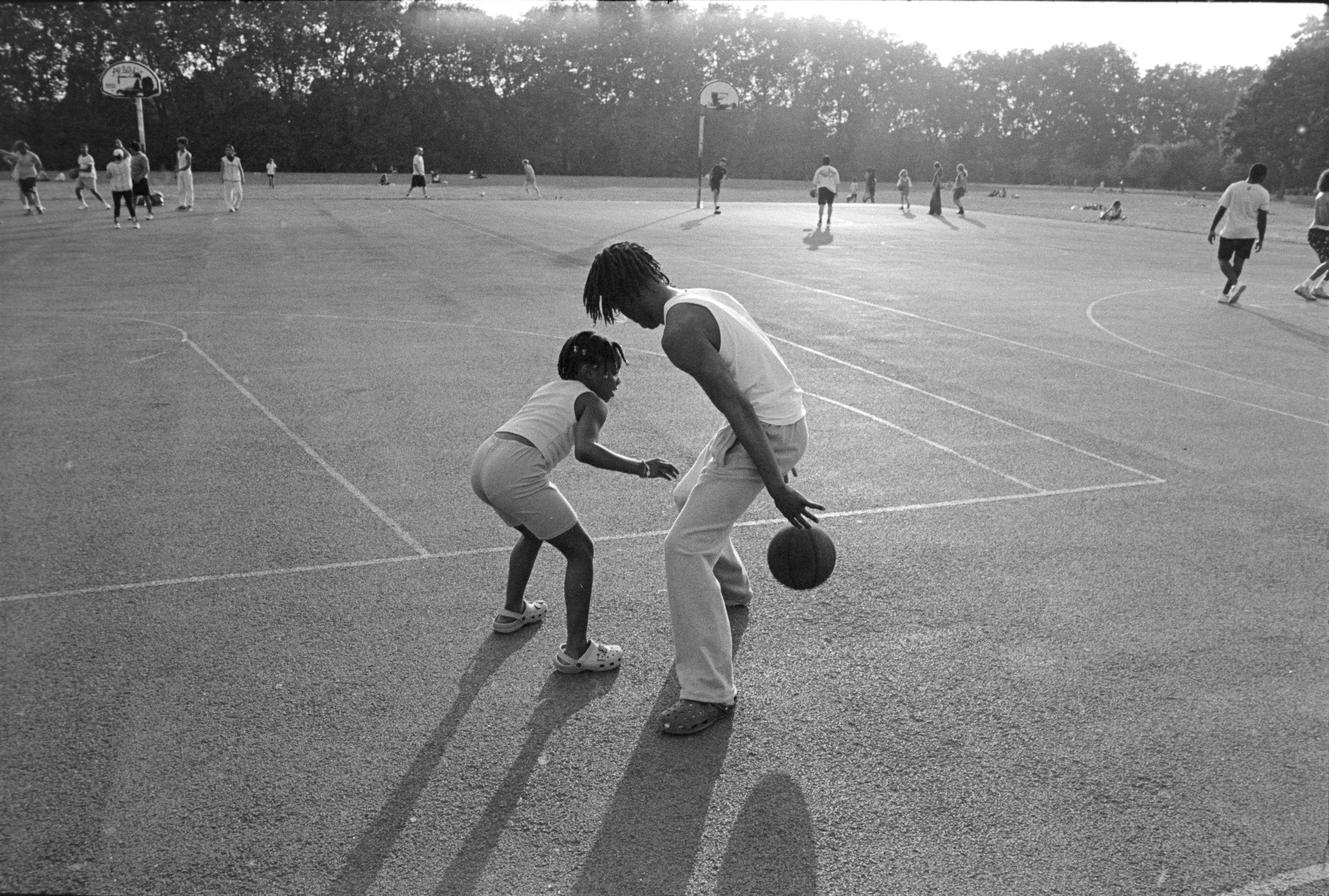 A man and a young girl playing basketball on an outdoor court during the daytime, with other people in the background. The man is holding a basketball, and the girl appears to be trying to guard him.