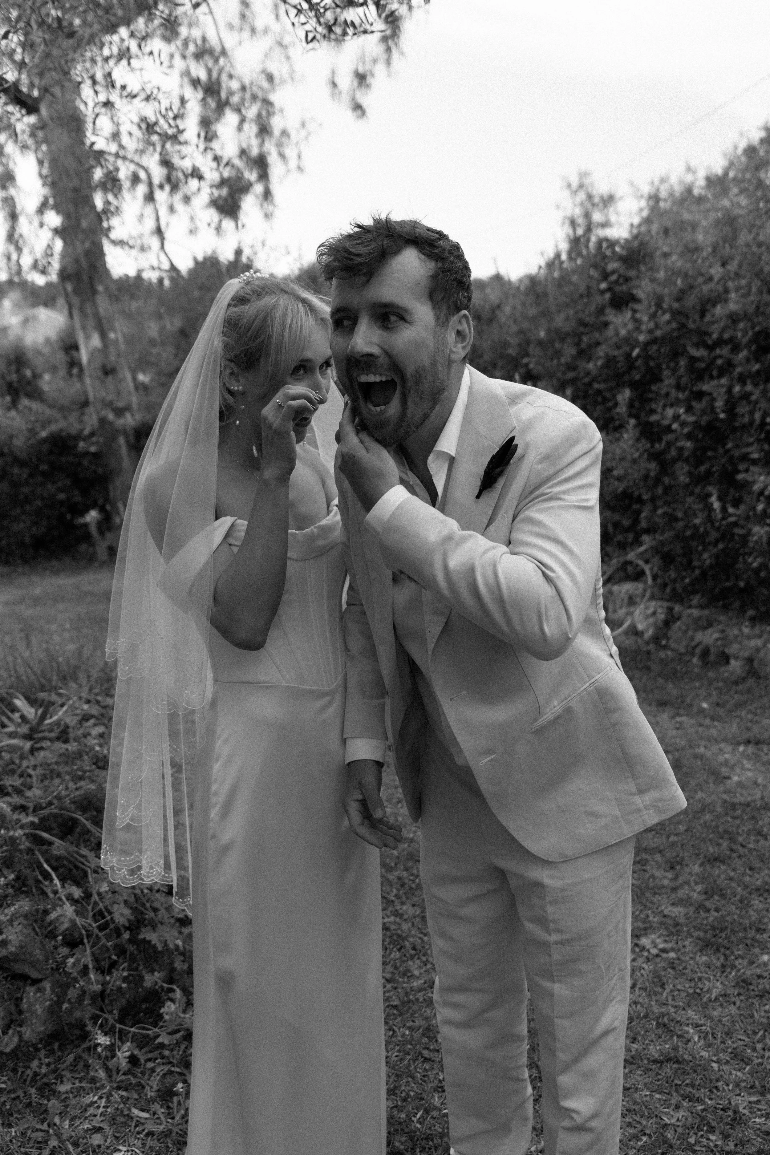 A black and white photo of a bride and groom outdoors, with the groom whispering into the bride's ear as she smiles and touches her face, surrounded by trees and bushes.