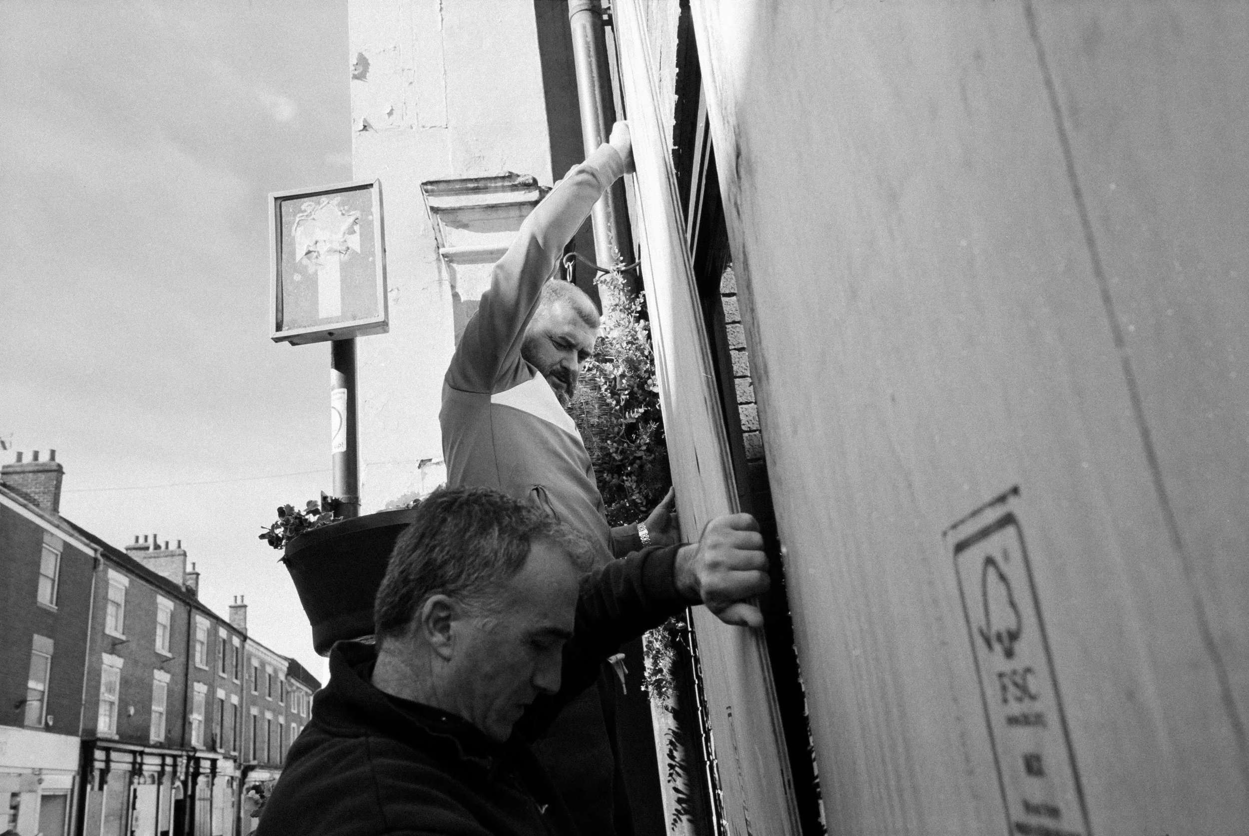 Two men are installing or working on a large wooden board or sign outside on a city street, with buildings visible in the background.