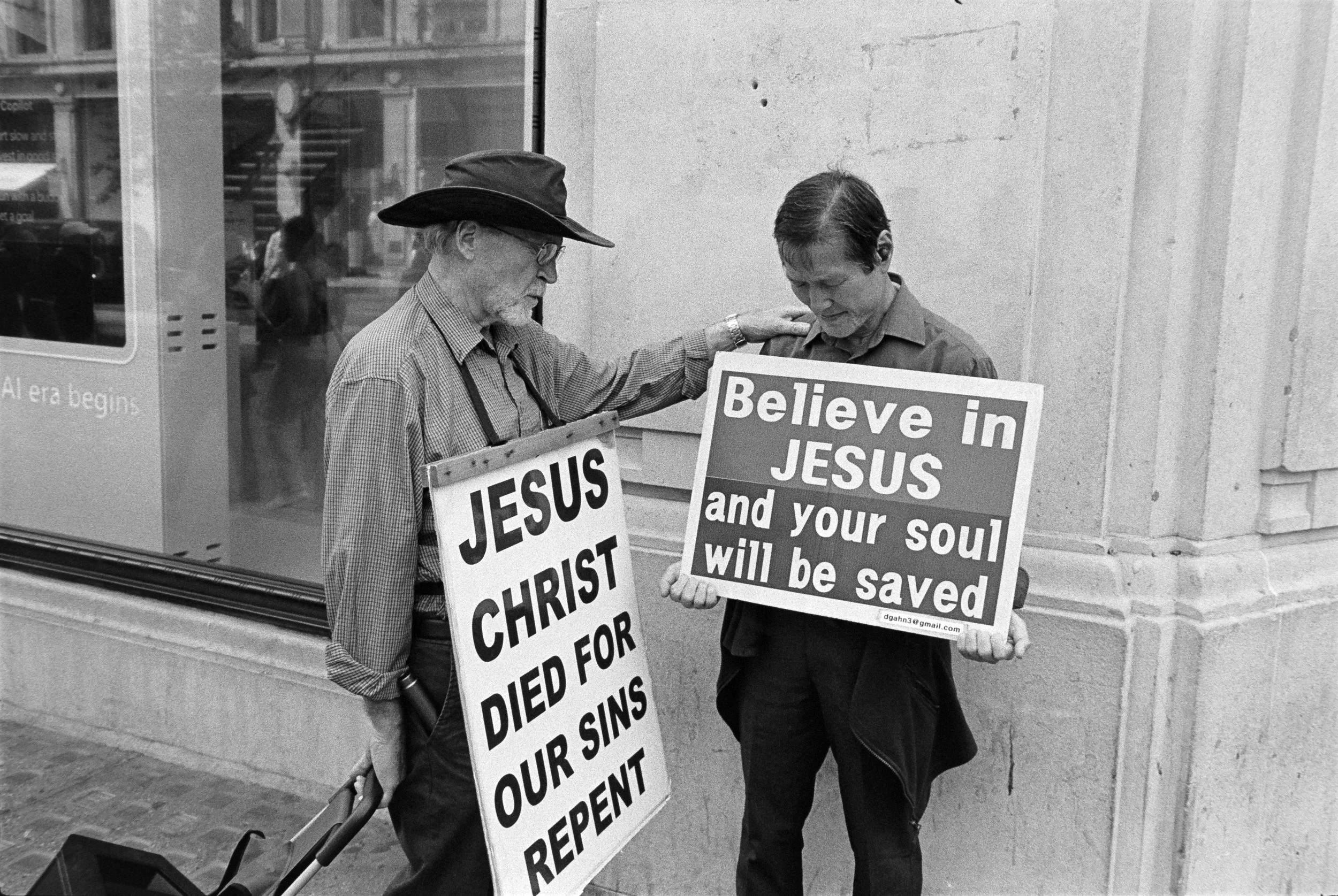 Two men standing on the sidewalk holding signs with religious messages; one sign reads "JESUS CHRIST DIED FOR OUR SINS REPENT," and the other reads "Believe in JESUS and your soul will be saved."
