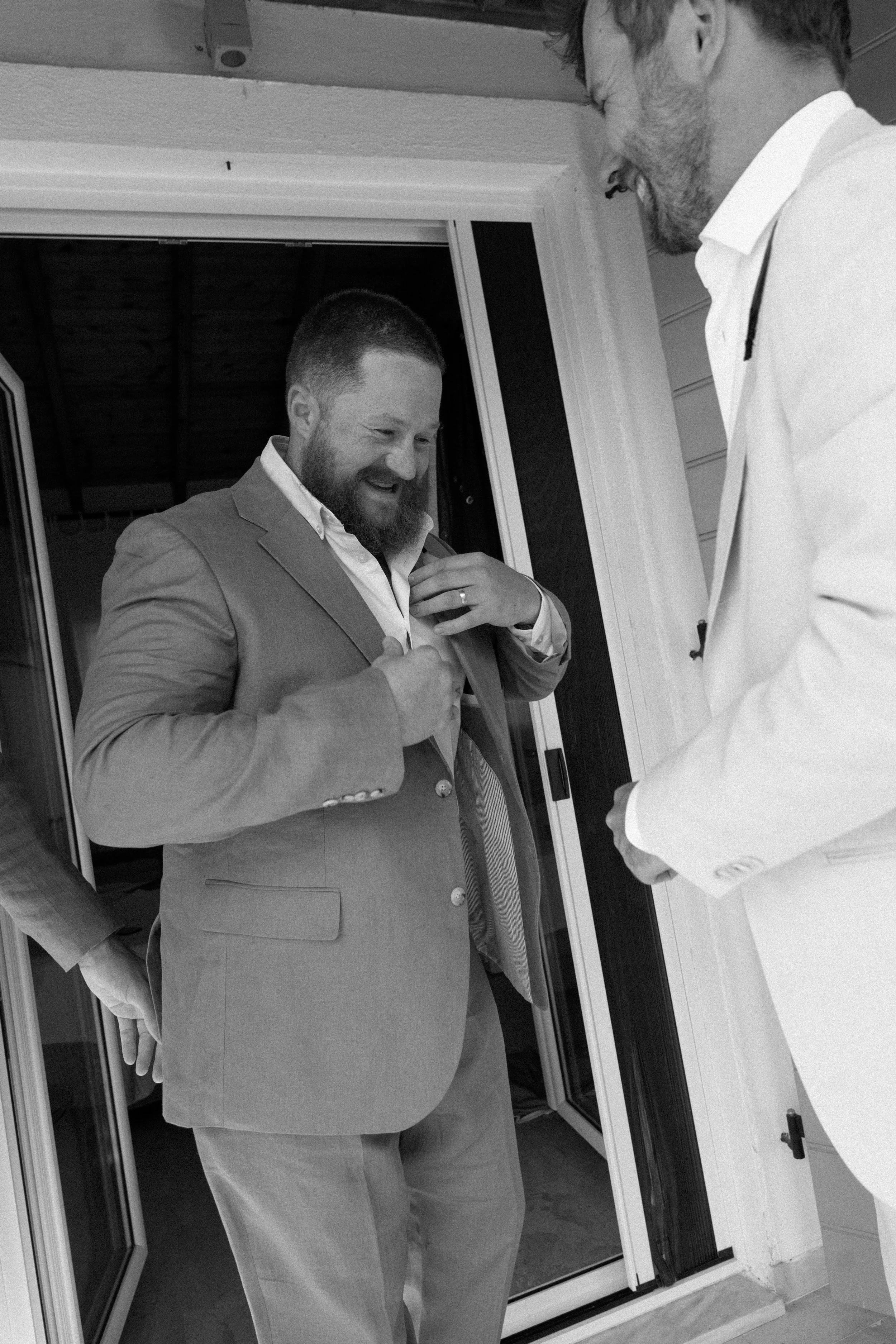 Two men in suits, one adjusting his jacket, share a laugh on a porch outside a house.