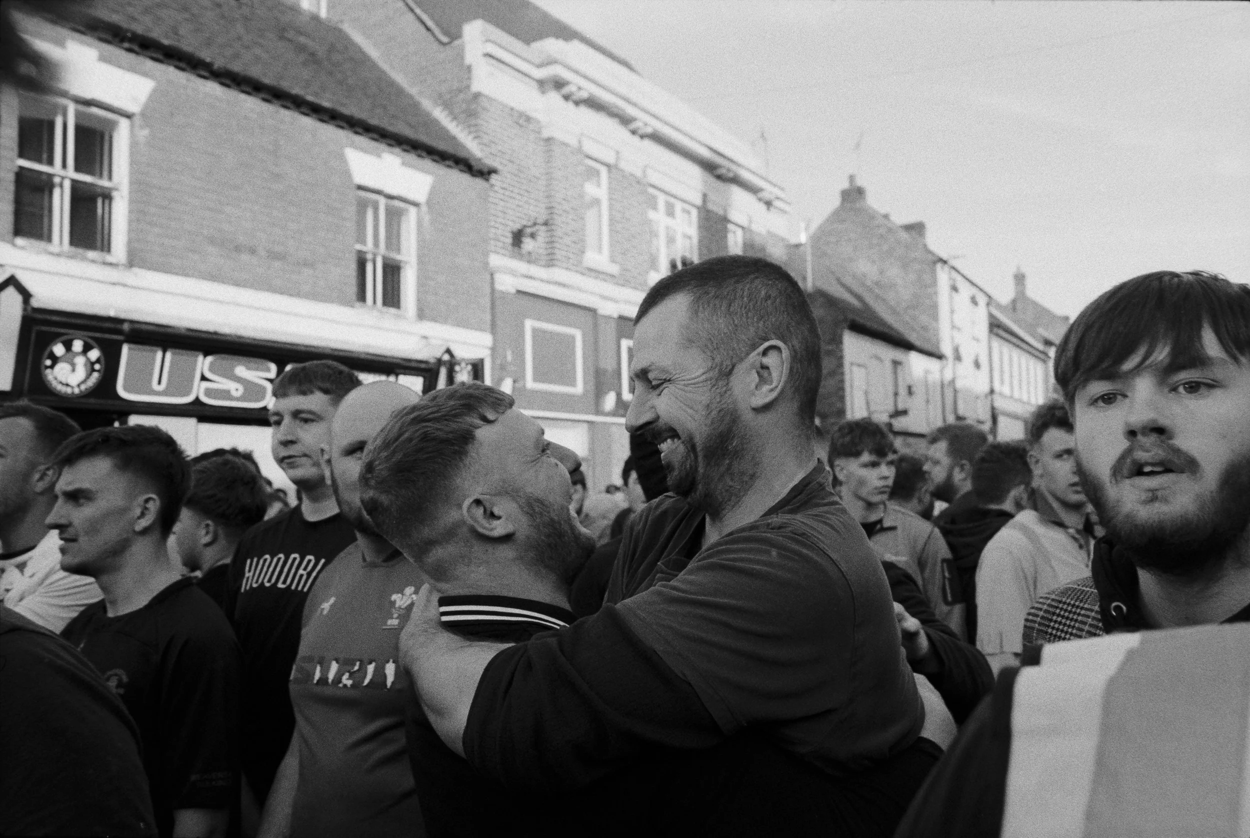 A group of people gathered outdoors, with two men in the center smiling and embracing each other.