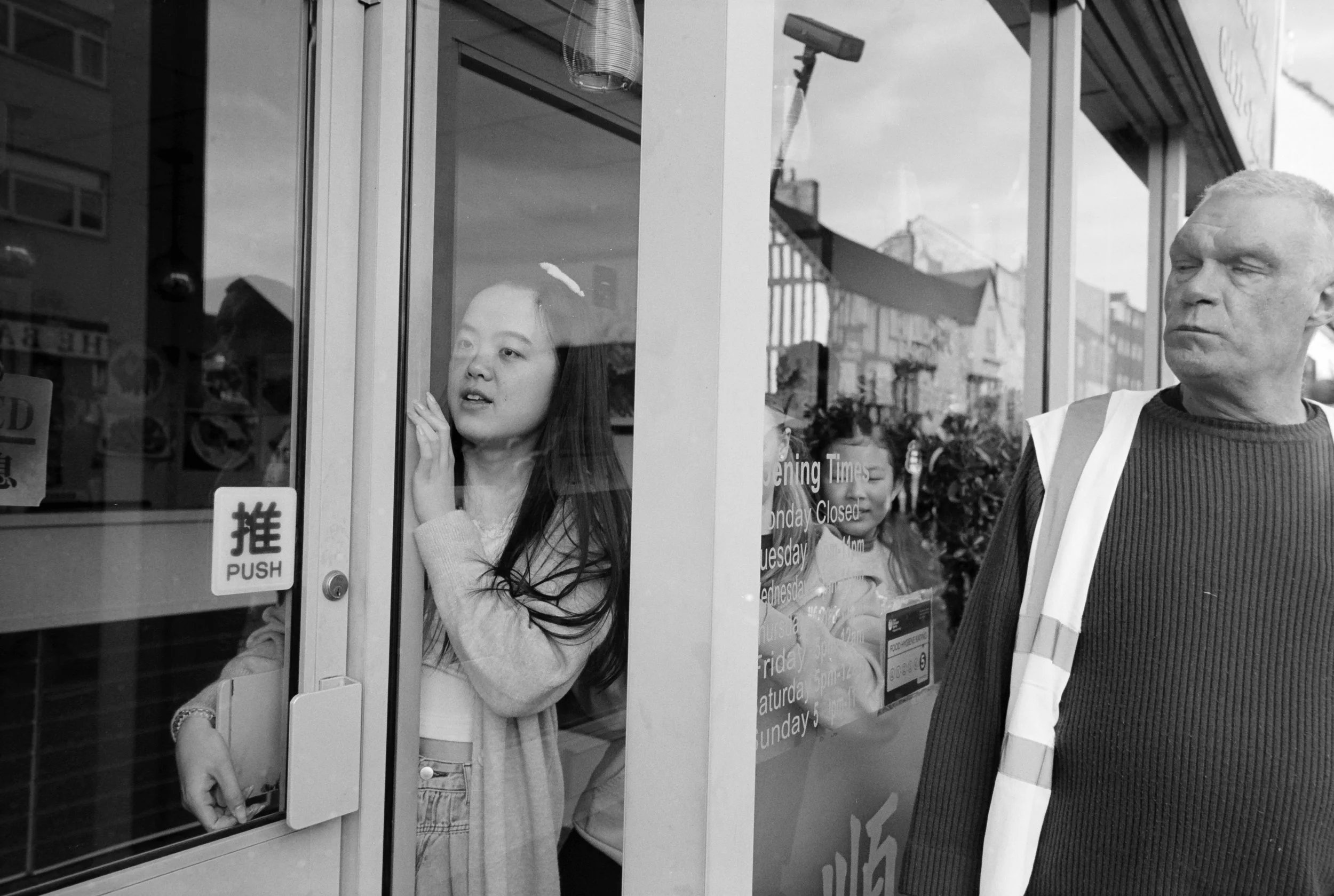 Three people outside a shop, one woman looking through the glass door, a second woman behind her, and a man standing to the right, wearing a reflective vest.