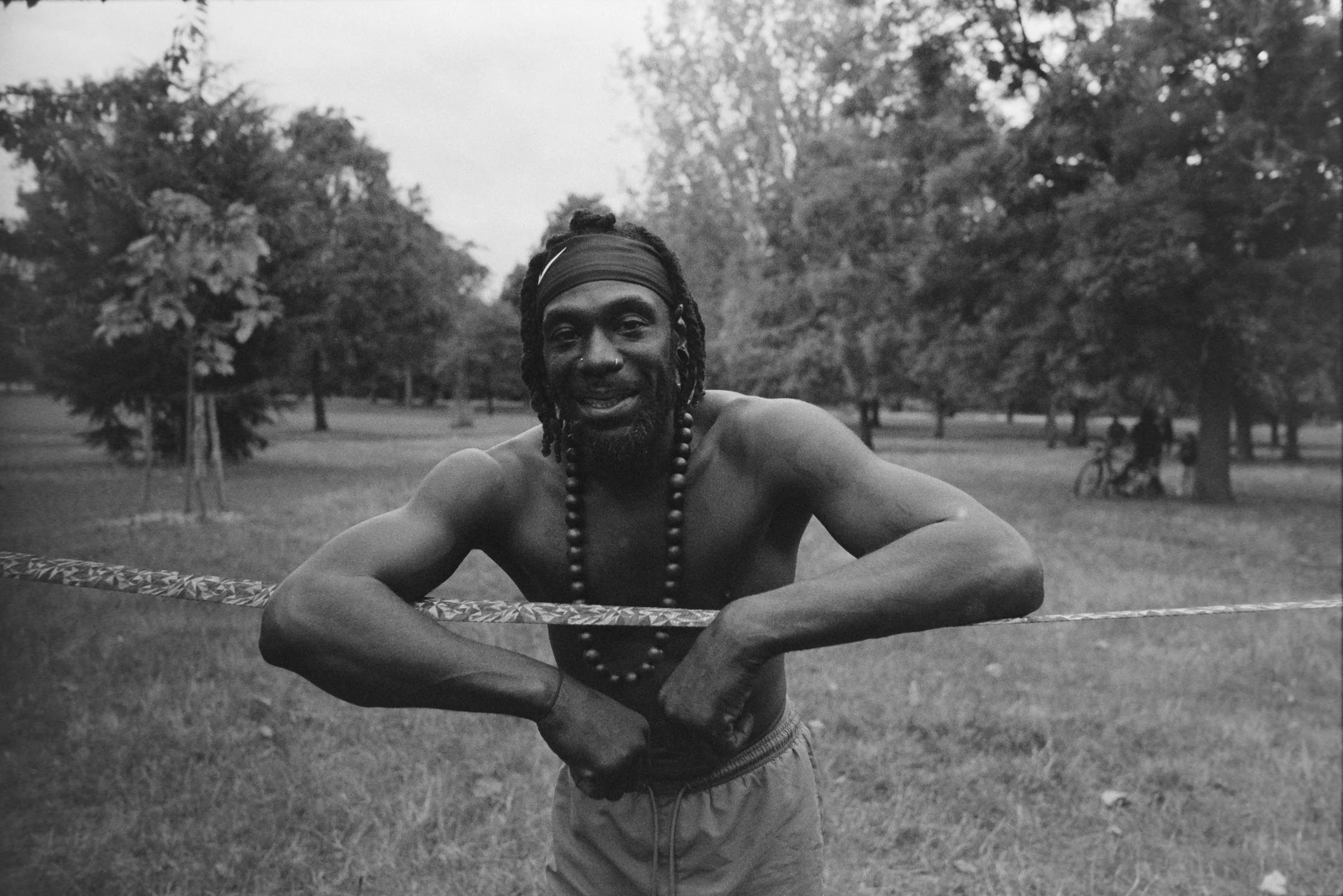 A shirtless man with dreadlocks and necklace standing in a park, holding a slackline, with trees and two cyclists in the background.