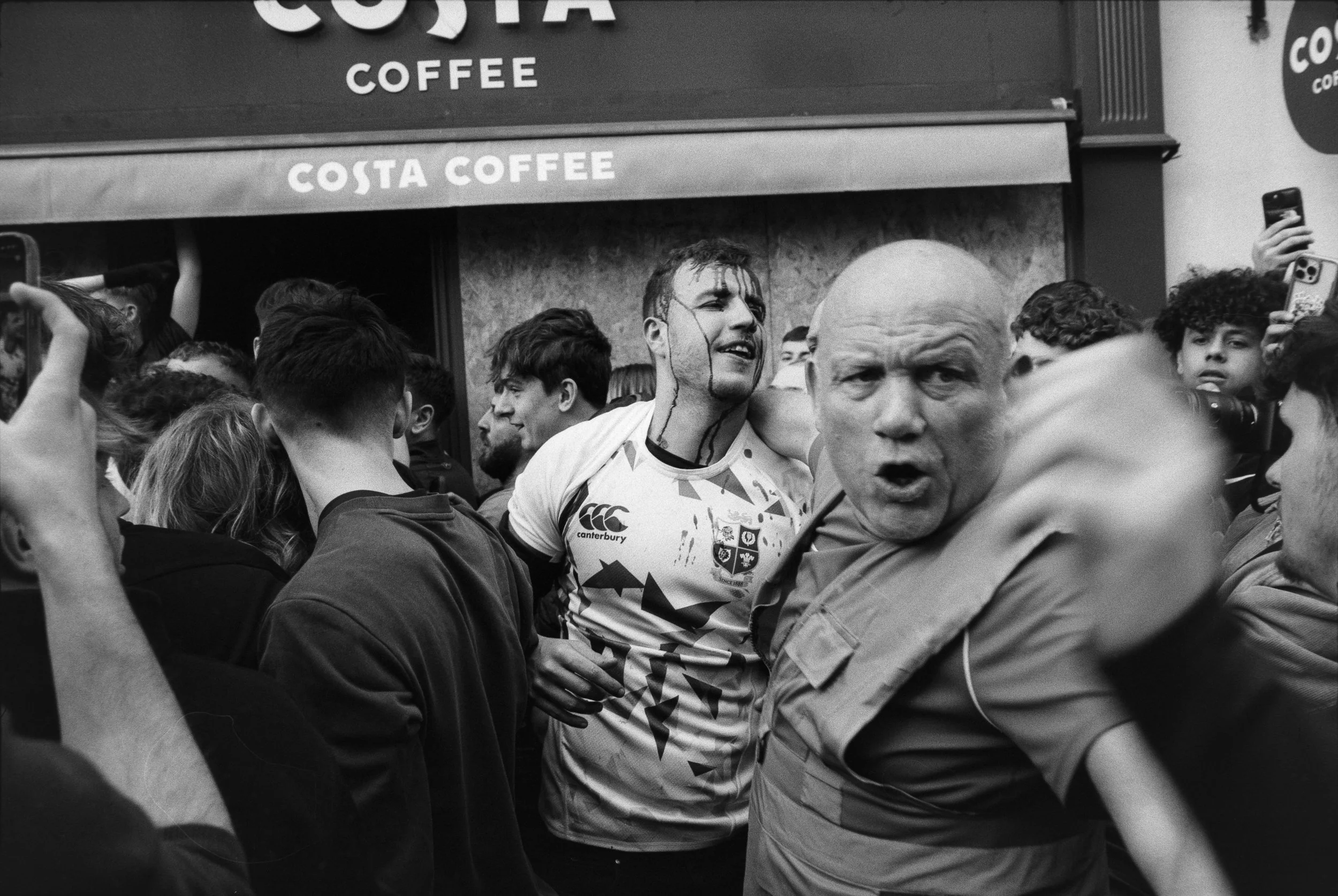 A crowd of people gathered outside a Costa Coffee shop, with two men in the foreground—one in a rugby jersey looking upset and another older man with an angry expression, reaching out with his hand.